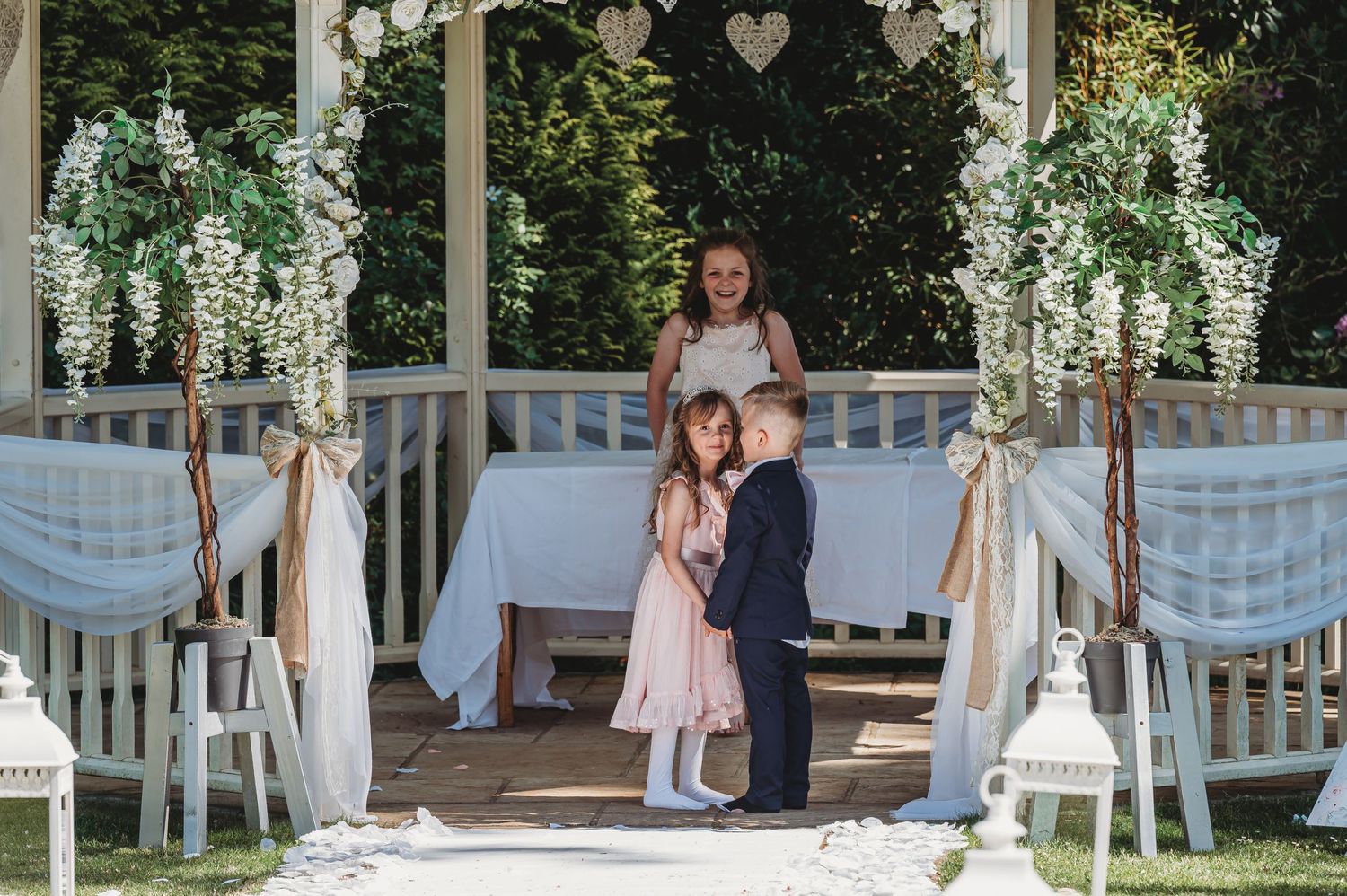 A beautifully decorated outdoor gazebo with white draping, floral arrangements and a wedding ceremony setup in a garden setting.