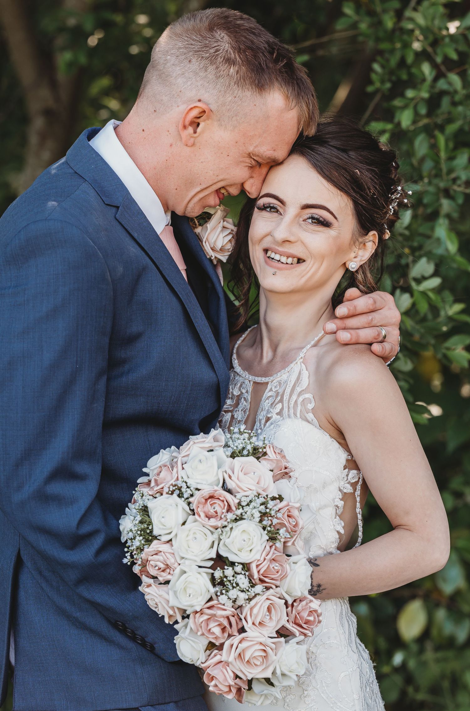 A joyous wedding couple embraces outdoors with a beautiful blush and white rose bouquet against green foliage.