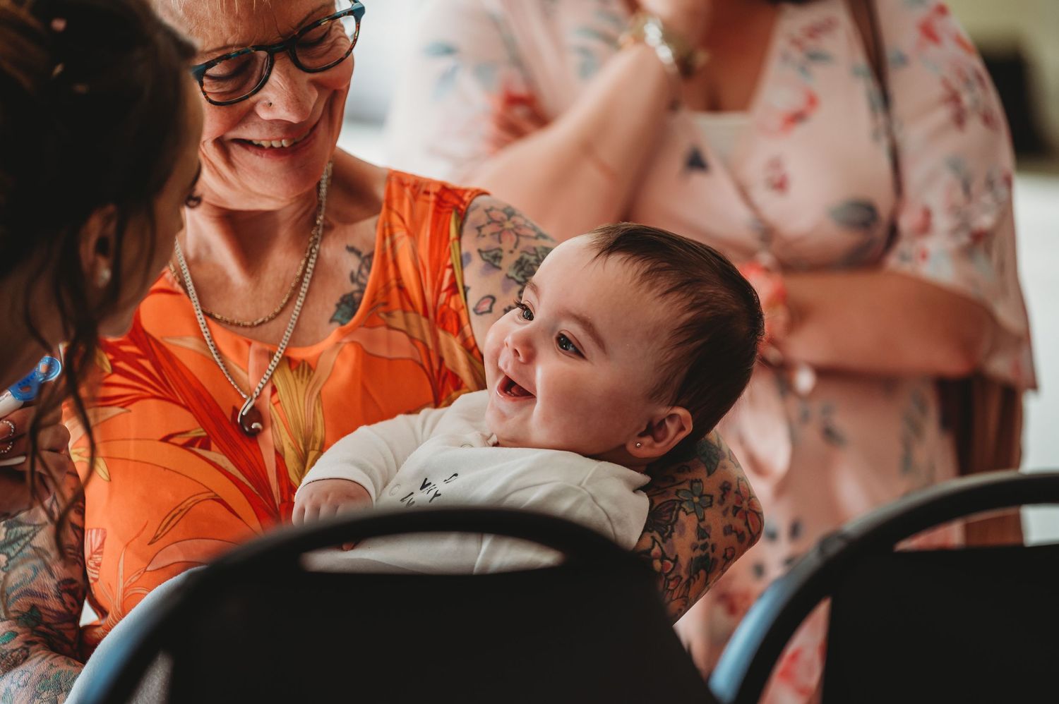 A happy child in a barber's chair laughs while getting a haircut as someone in a bright orange shirt looks on.