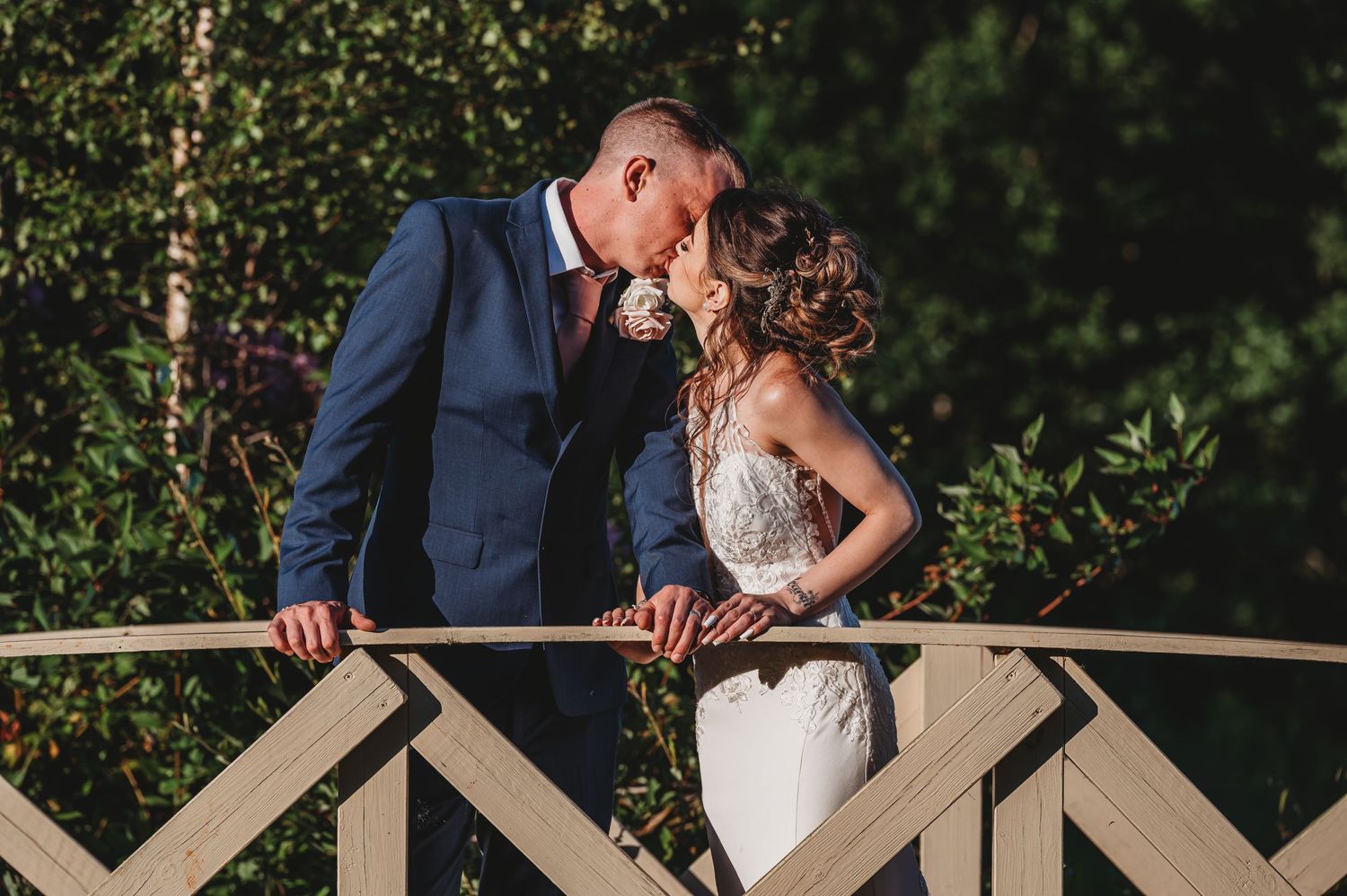 Newlyweds share a romantic moment while leaning on a wooden bridge railing in an outdoor garden setting.