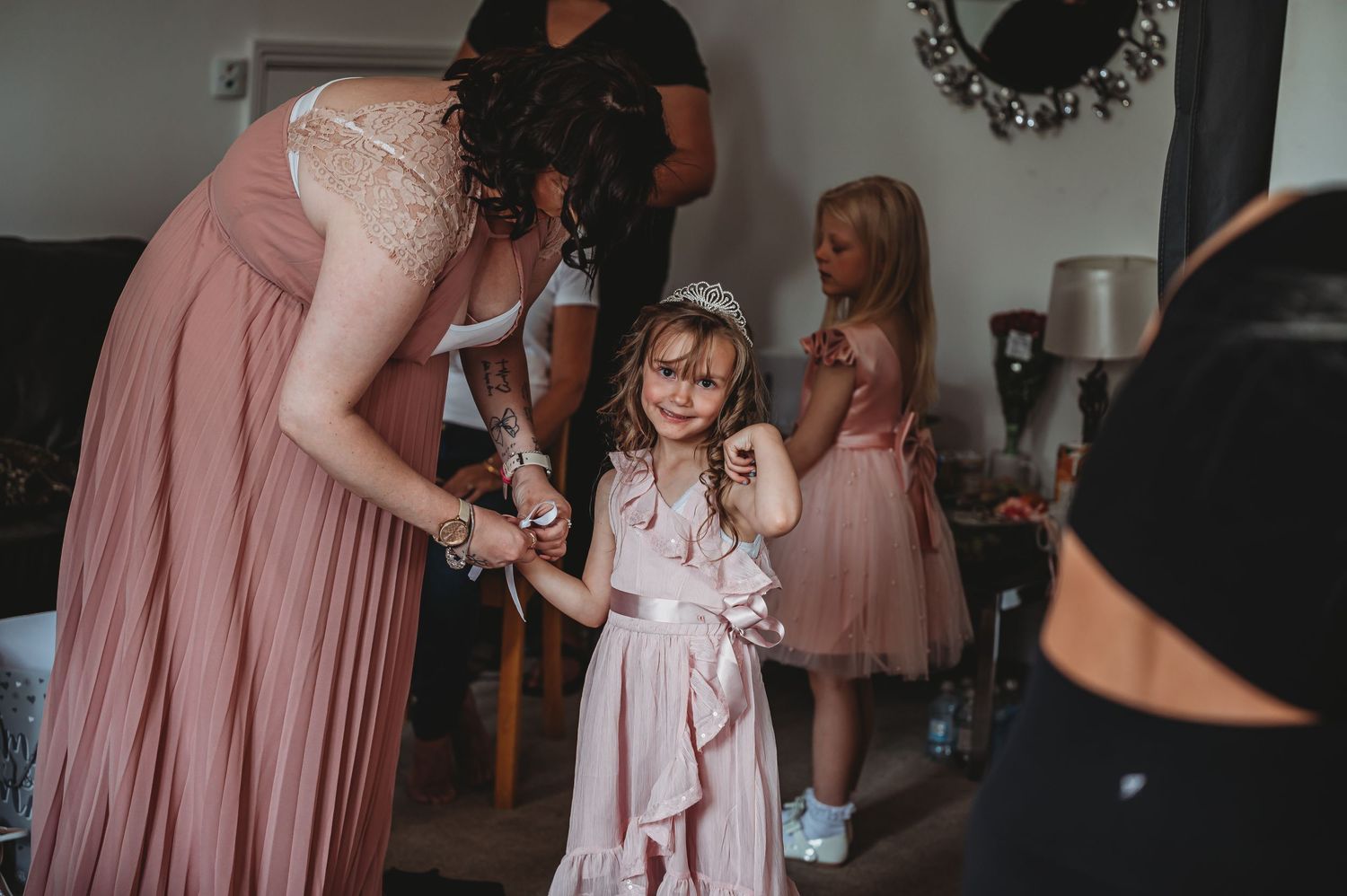 People gather indoors dressed in pink formal attire while helping a young girl with her outfit.