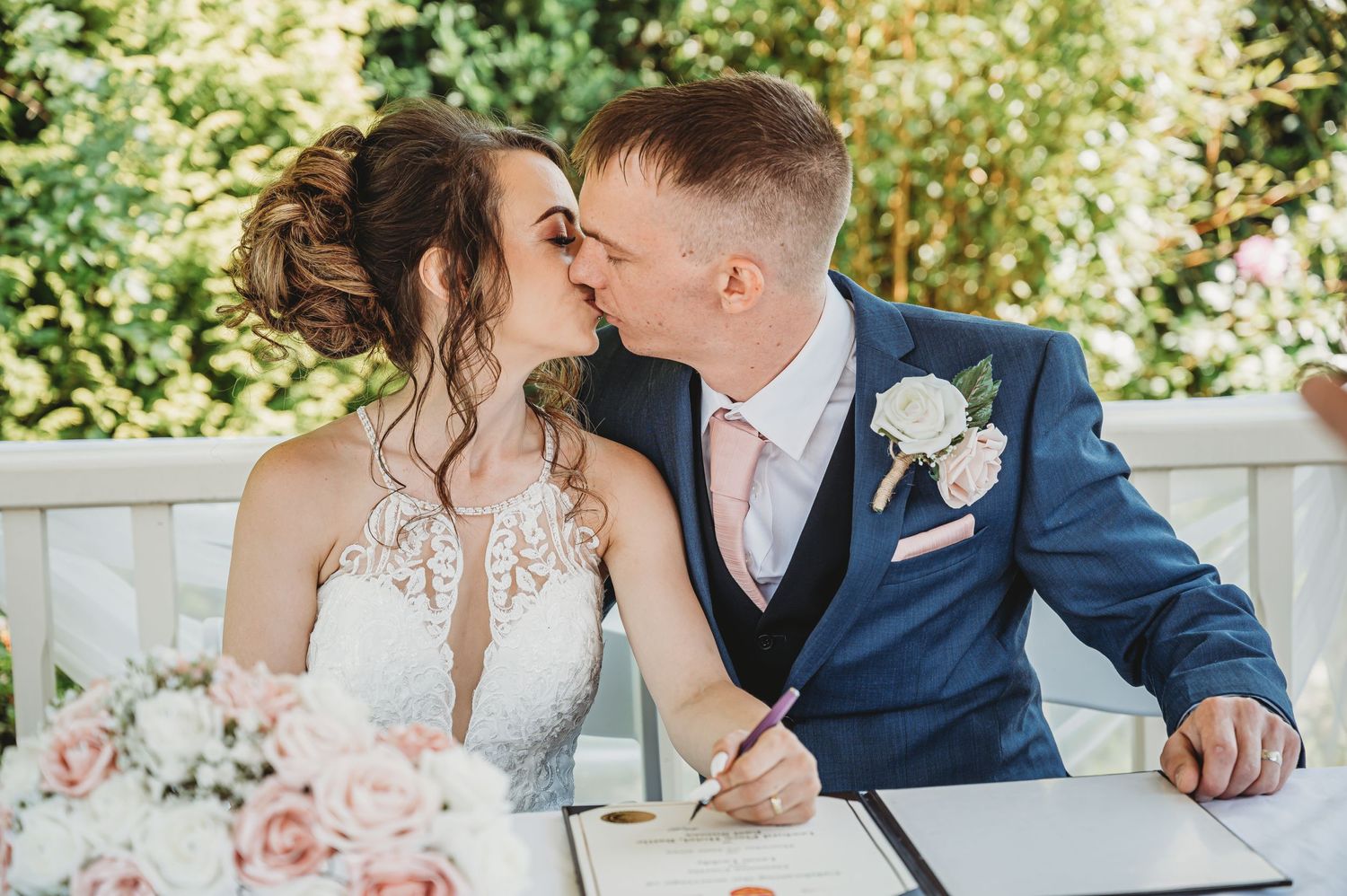 A couple shares a romantic kiss while signing marriage documents at an outdoor ceremony with pink floral decor.