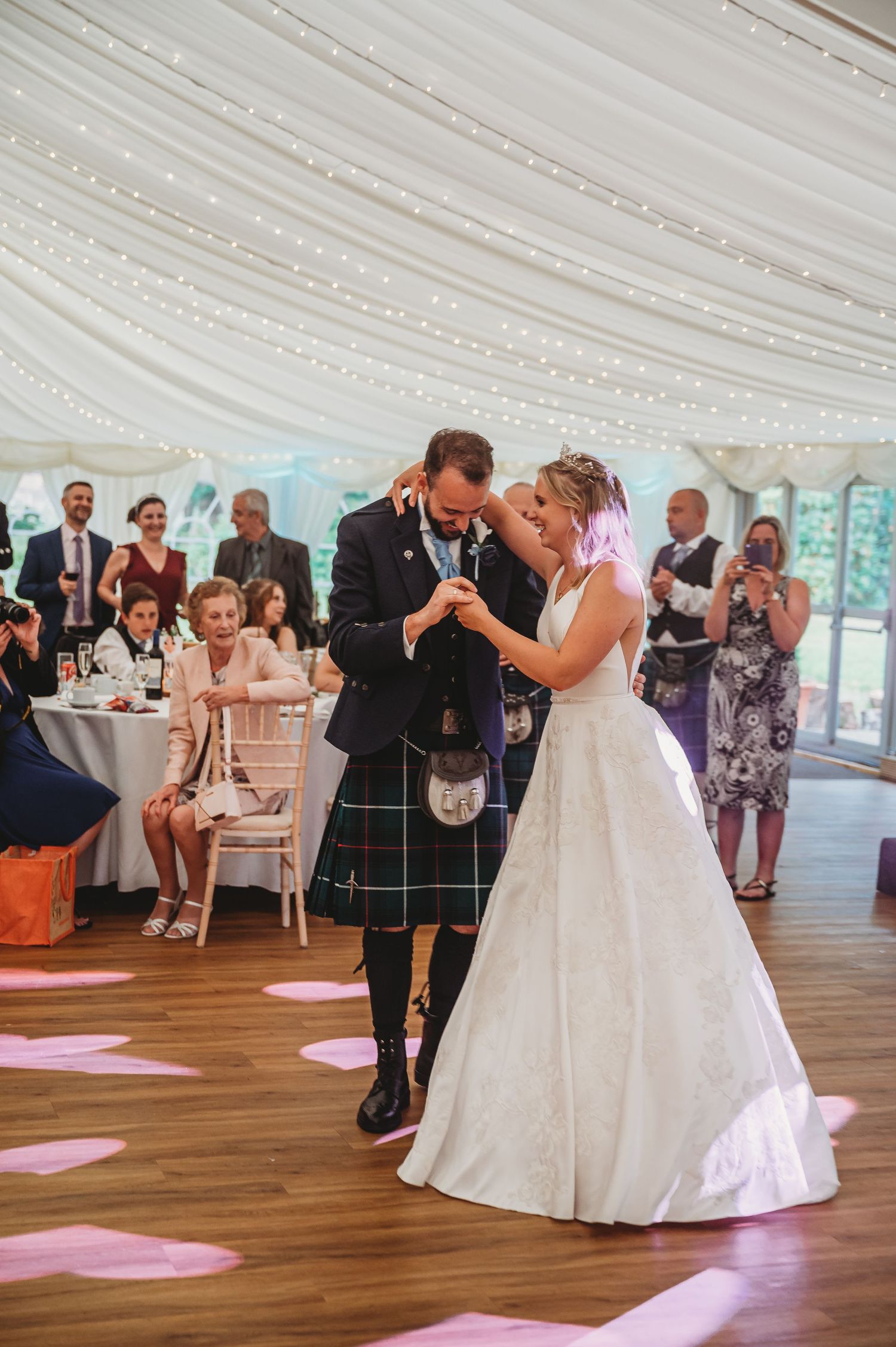 Couple shares their first dance at a Scottish wedding celebration under a draped tent ceiling with guests watching.