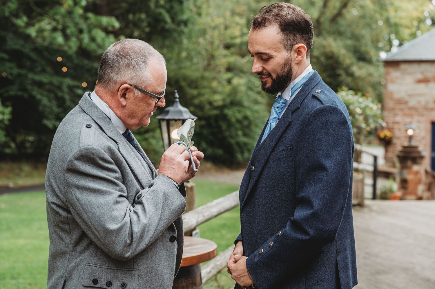 Two people in suits share a moment outdoors near a stone building, one lighting a cigarette for the other.