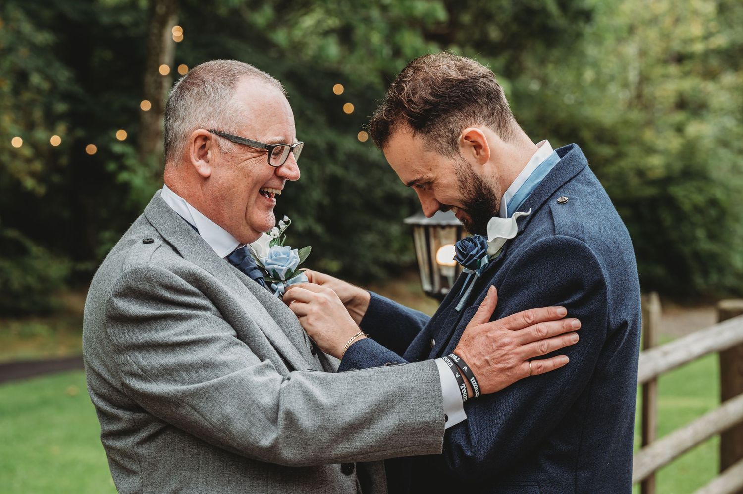 Two men share an emotional moment while adjusting boutonnieres at an outdoor wedding ceremony.