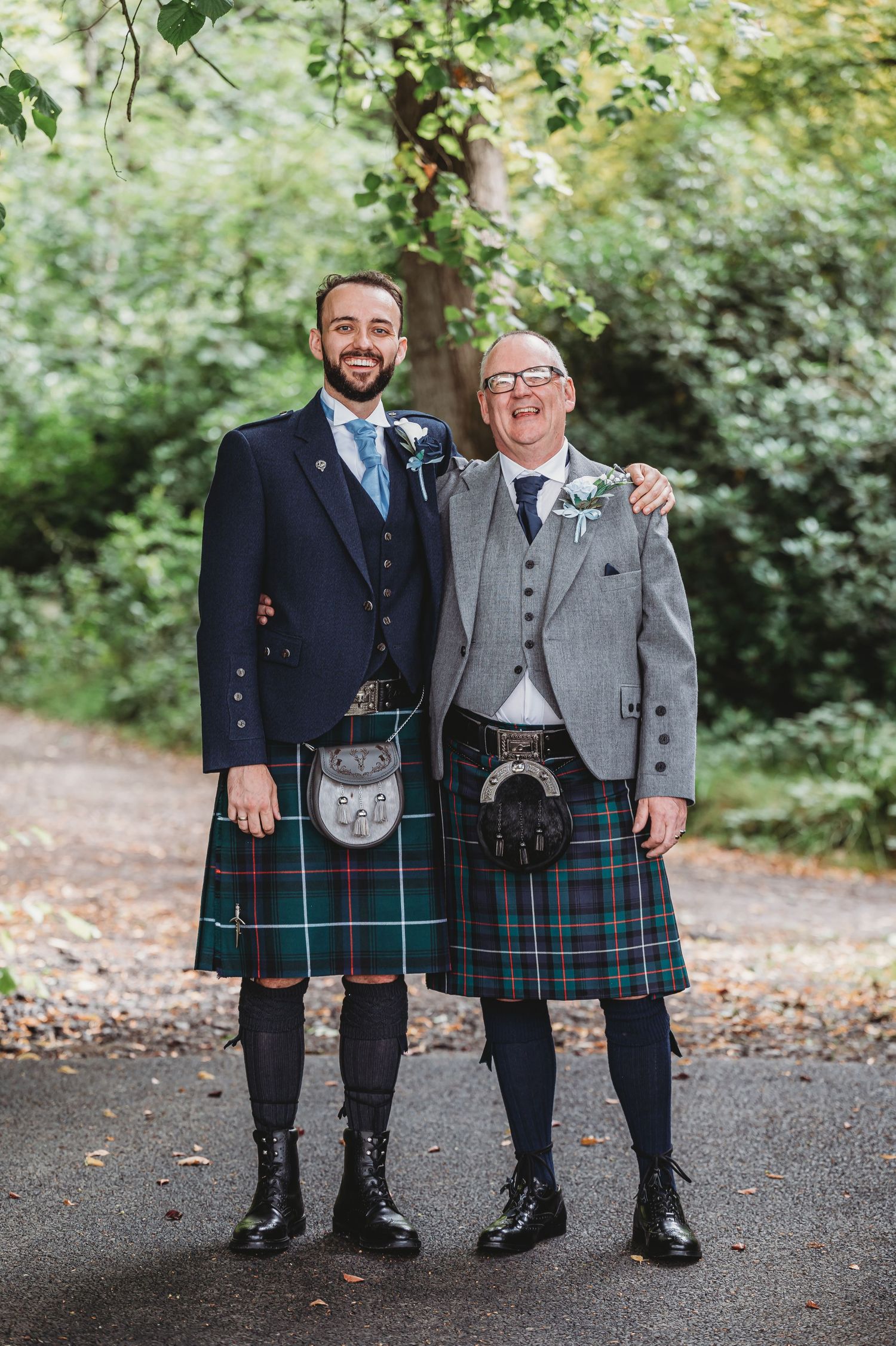 Two men in traditional Scottish kilts and jackets pose on a forest path during a wedding celebration.