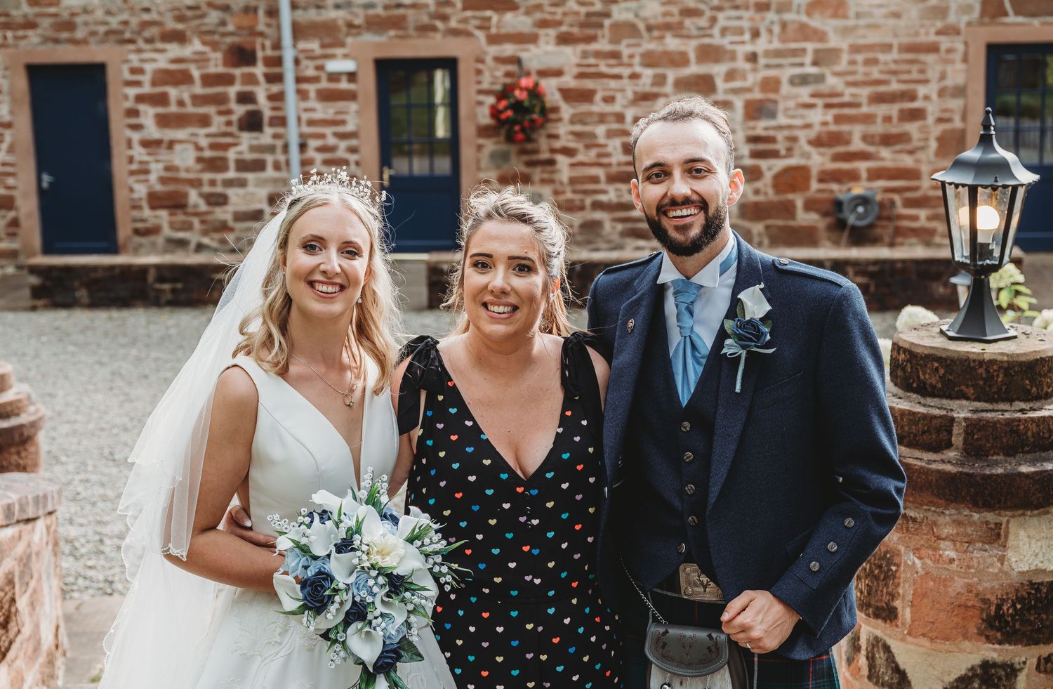 Three people smile together at a rustic brick venue, with a bride in white holding a blue and white bouquet.
