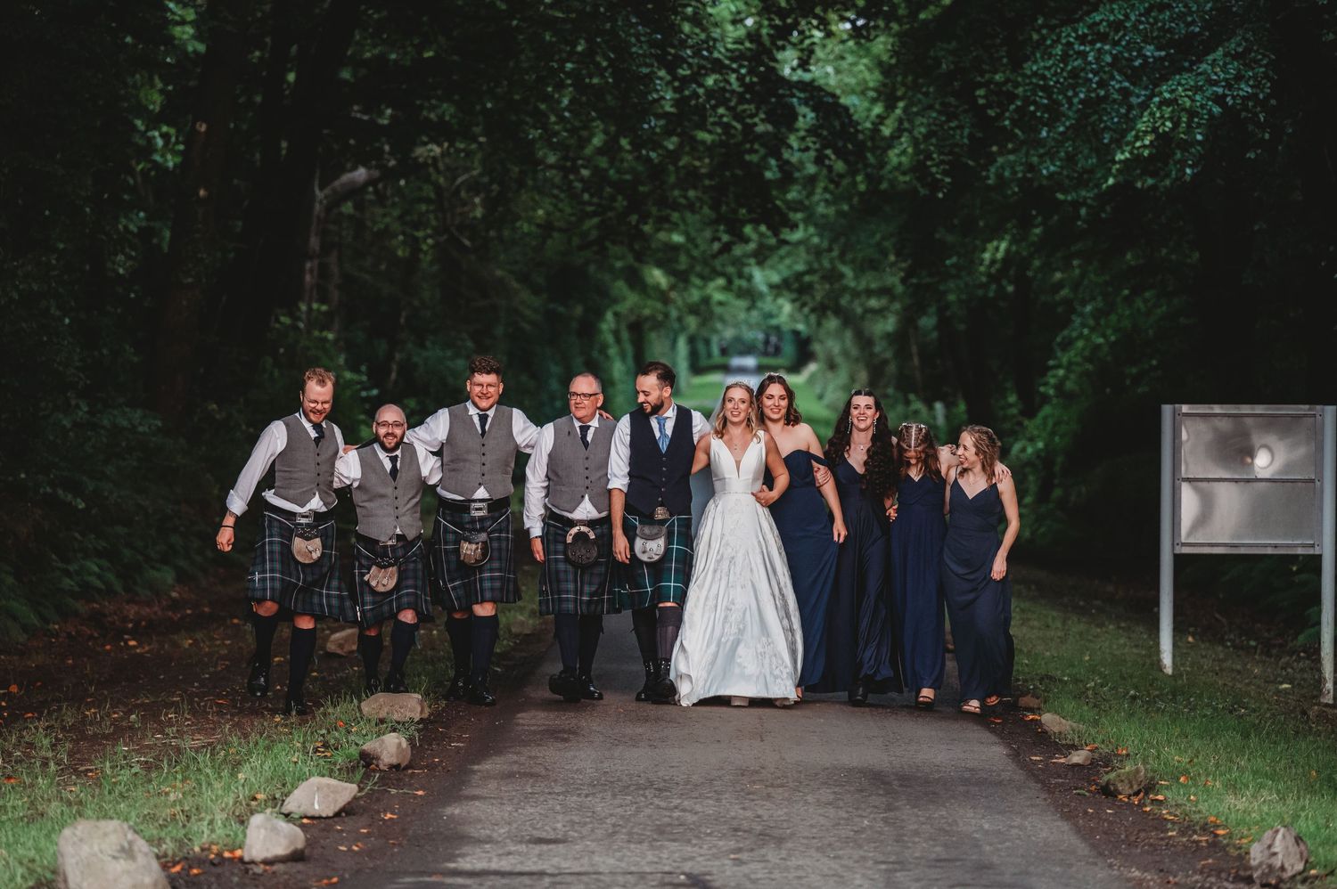 Wedding party in kilts and formal dresses poses together on a tree-lined path outdoors.