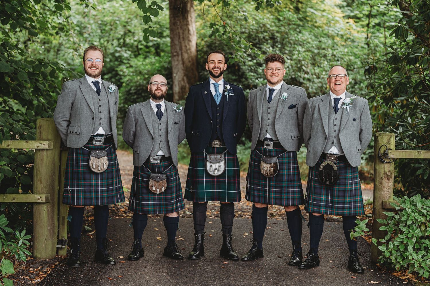 Five men in traditional Scottish kilts and formal attire stand together on a wooded path.
