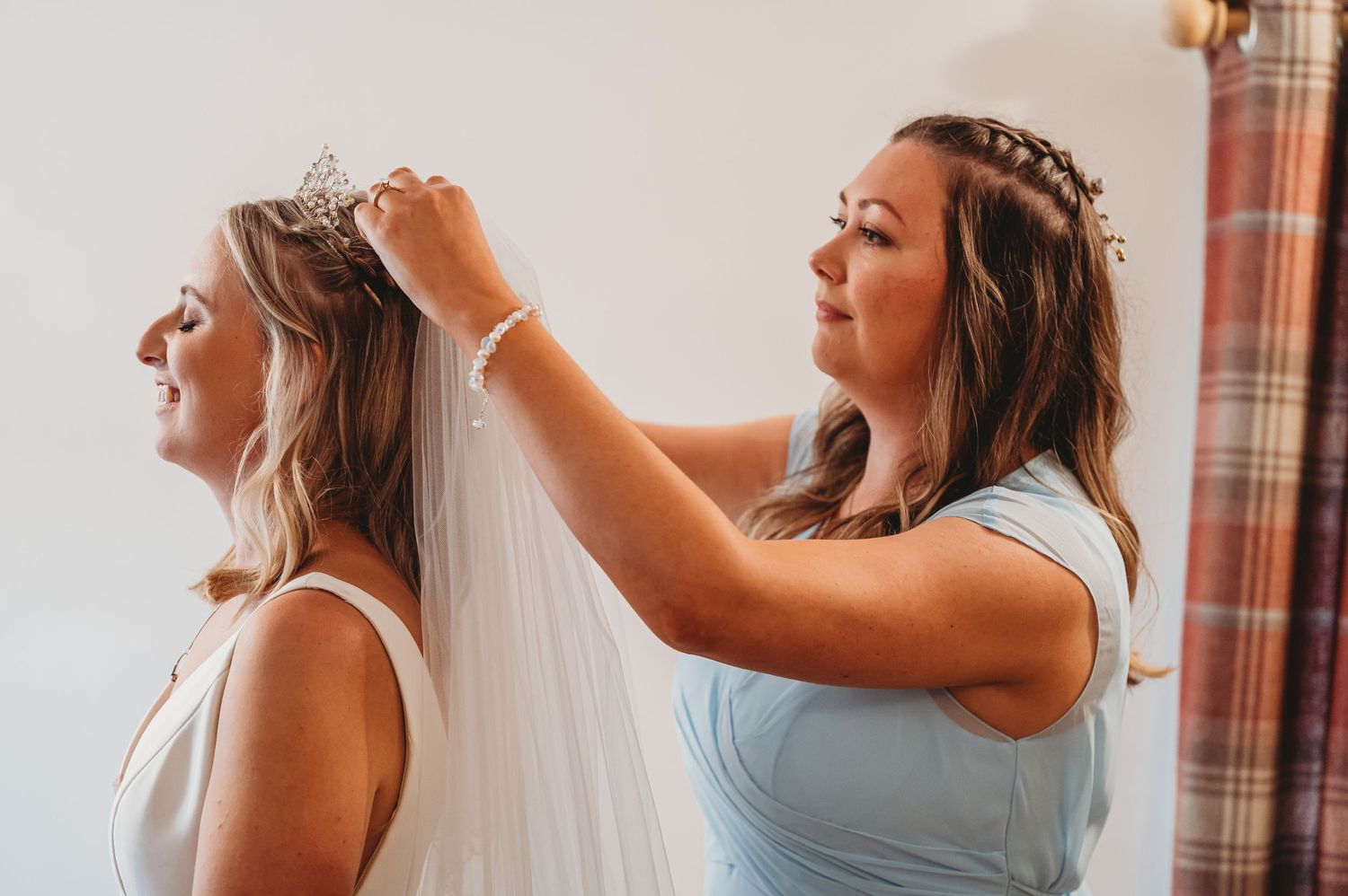A helping hand adjusts a flowing white wedding veil during wedding preparations in warm natural lighting.