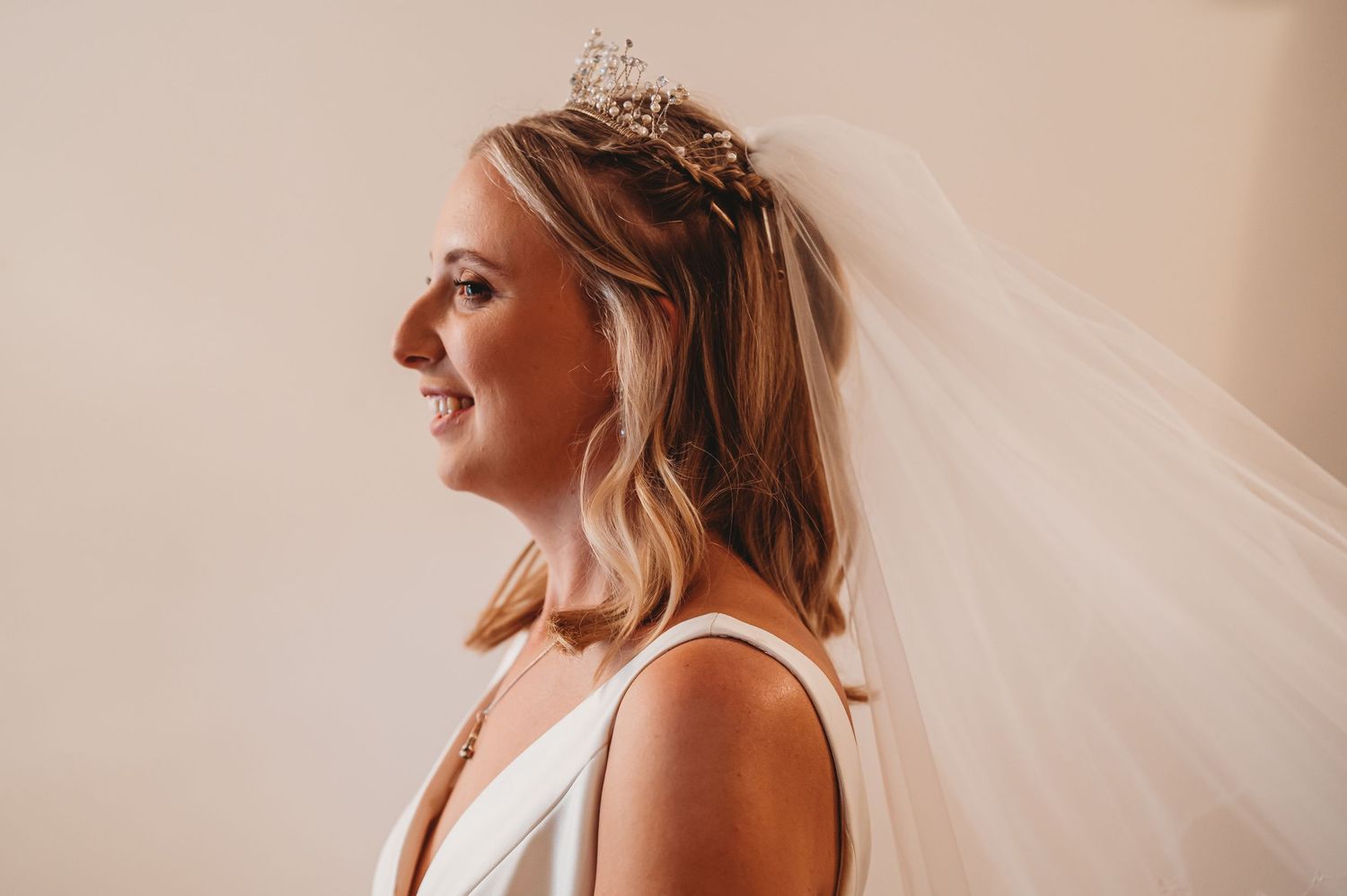 A bride in profile view wearing a white sleeveless dress, delicate tiara and flowing veil against a neutral background.