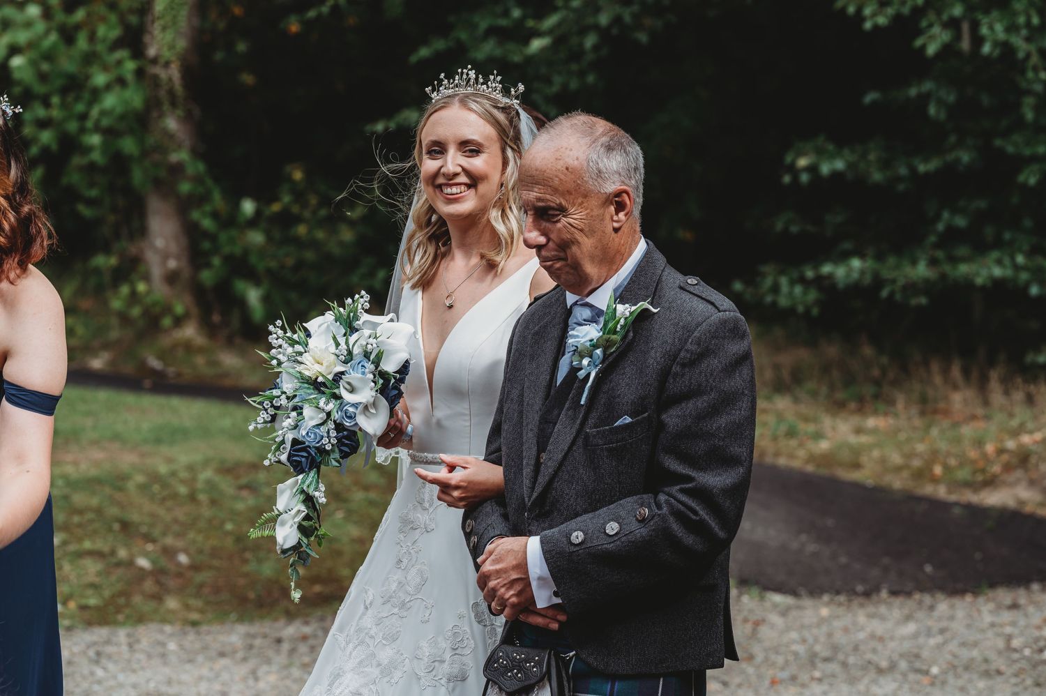 A bride in a white lace gown holding a blue and white bouquet walks arm in arm with an older man in a suit.