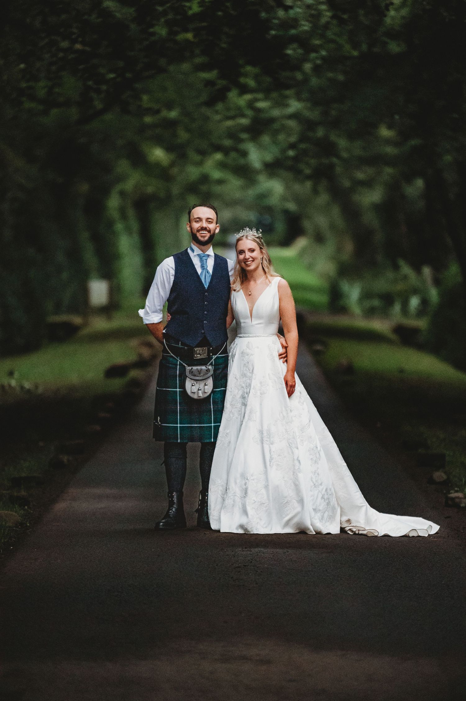 A bride in a white lace gown and groom wearing a traditional kilt pose together on a tree-lined path.