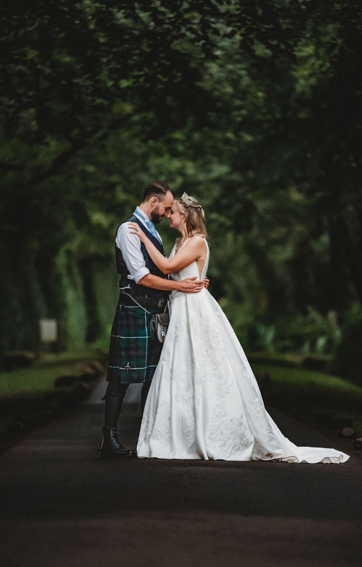 A romantic wedding couple embrace on a tree-lined path, with the groom in traditional Scottish kilt and bride in flowing white gown.