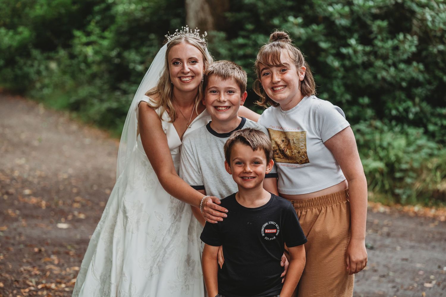 A group portrait on a forest path with someone wearing a white wedding dress and three children gathered close together.