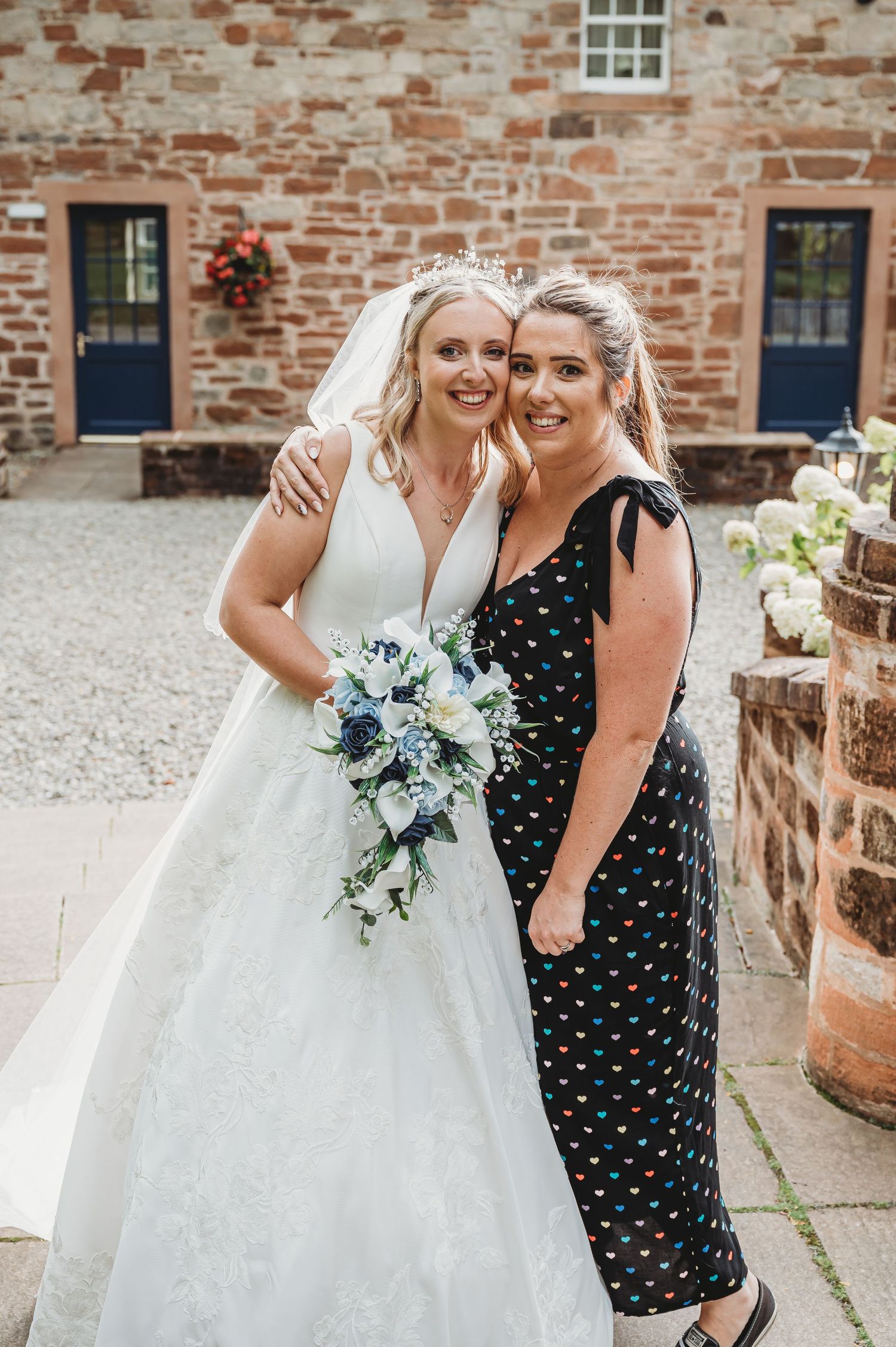 Two women, one in a white wedding gown and one in a floral dress, stand together in front of a stone wall.