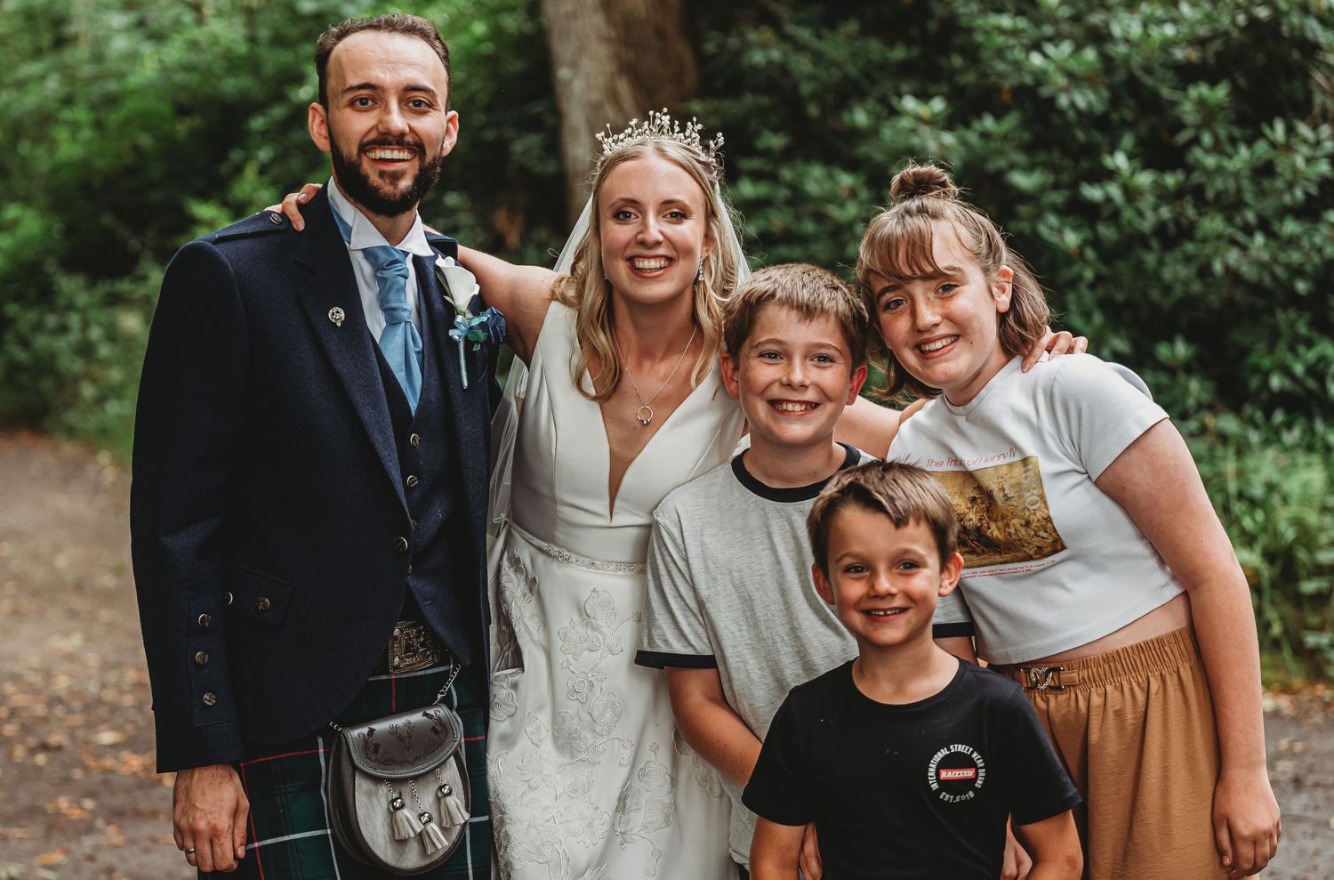 A joyful wedding photo showing a couple and three children posing together outdoors surrounded by green foliage.