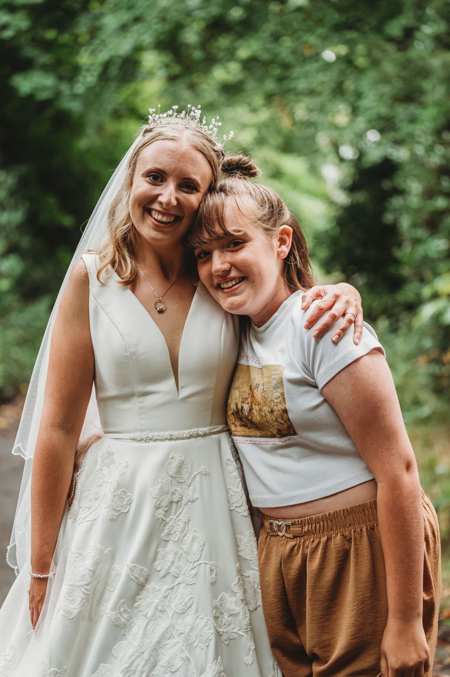 A bride in a white wedding dress with plunging neckline and veil poses with a guest in casual attire in an outdoor setting.