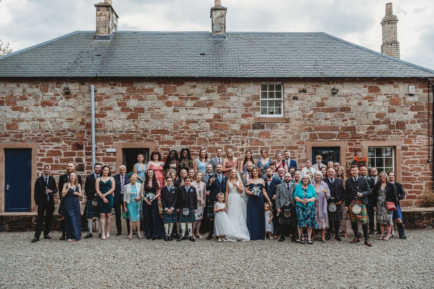 Wedding celebration group photo outside rustic stone building with slate roof and brick exterior.