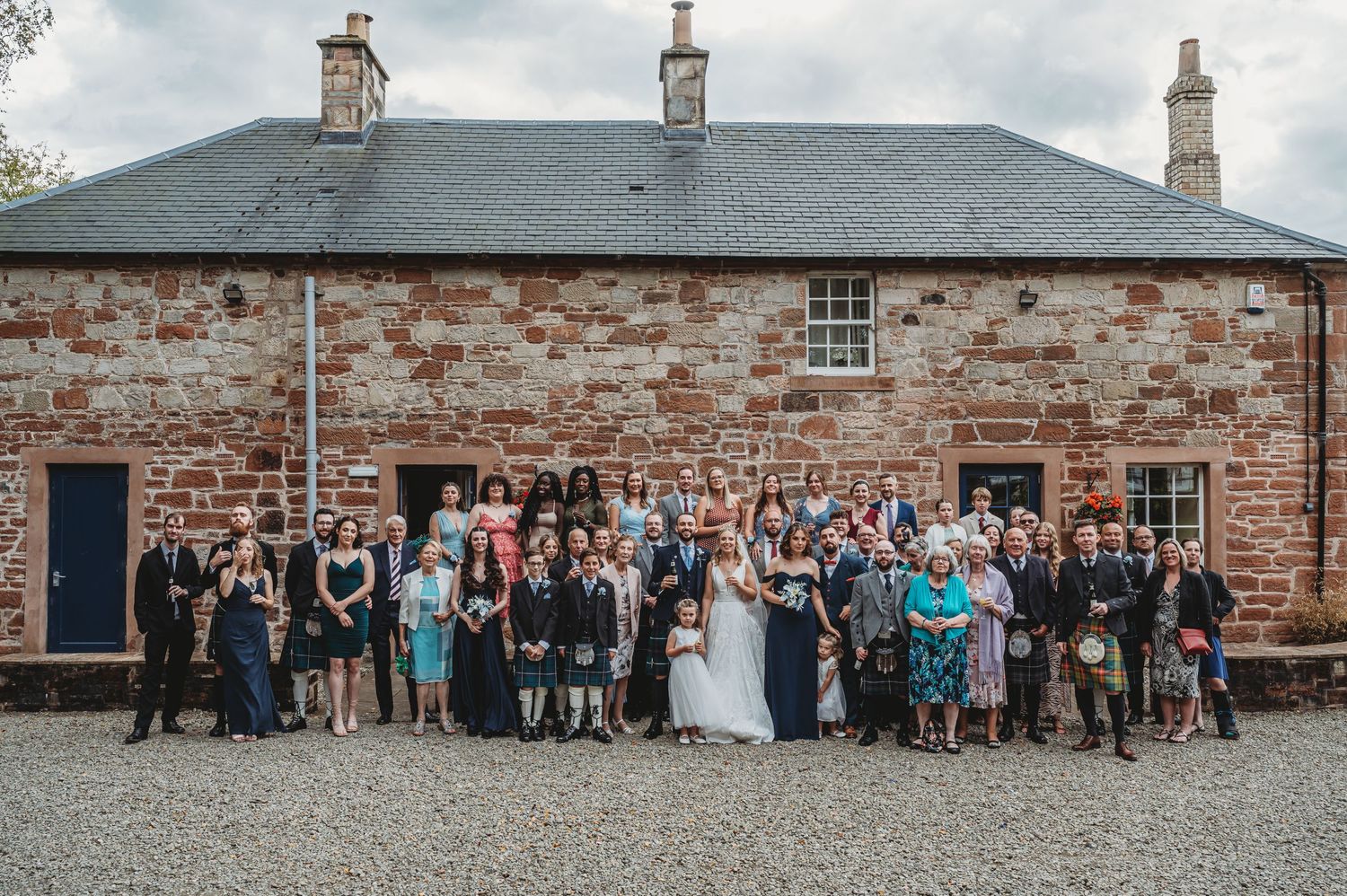 A large wedding party poses for a group photo in front of a rustic brick building with three chimneys.