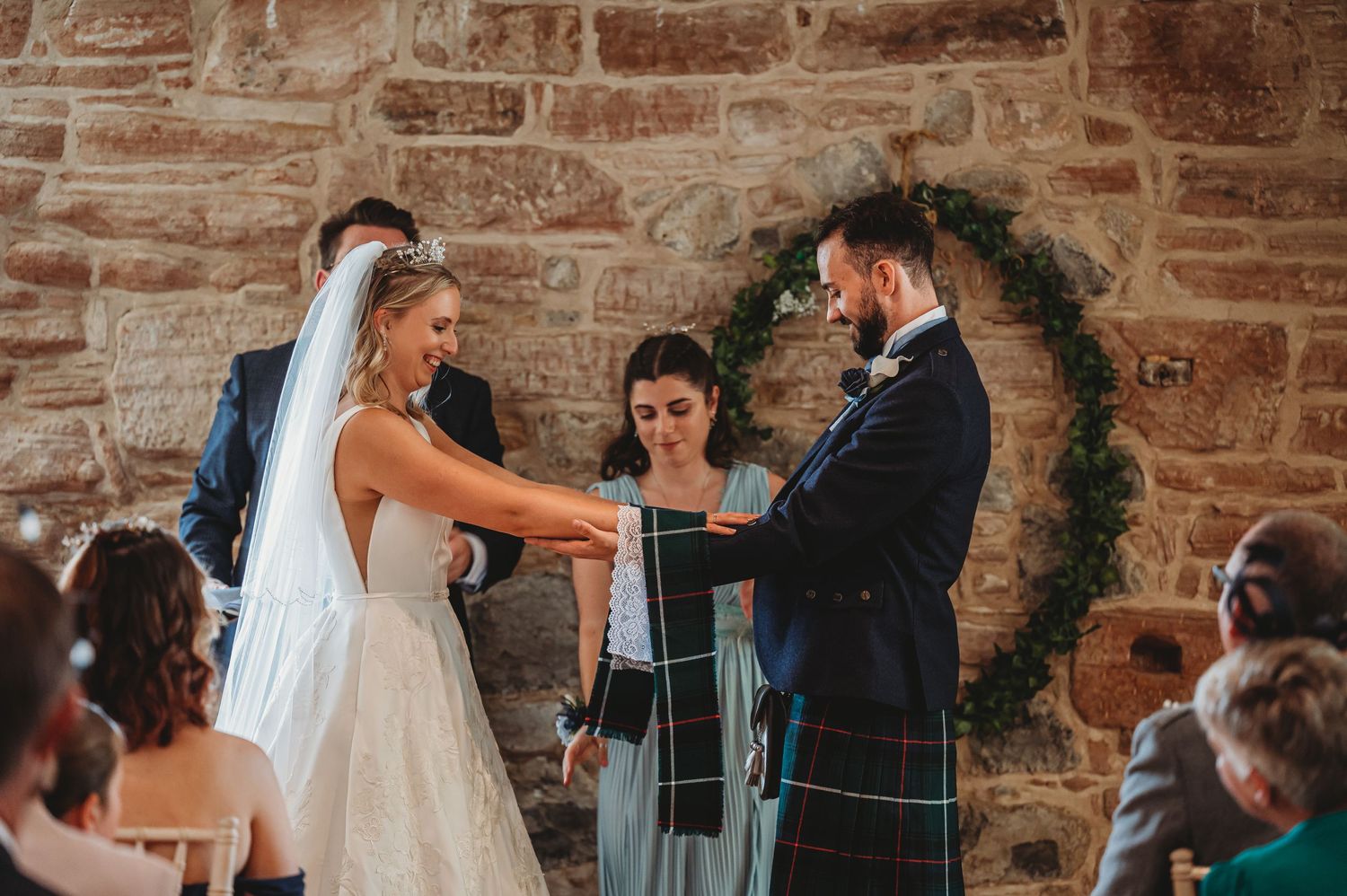 A wedding ceremony takes place in front of a stone wall under an evergreen arch with guests looking on.