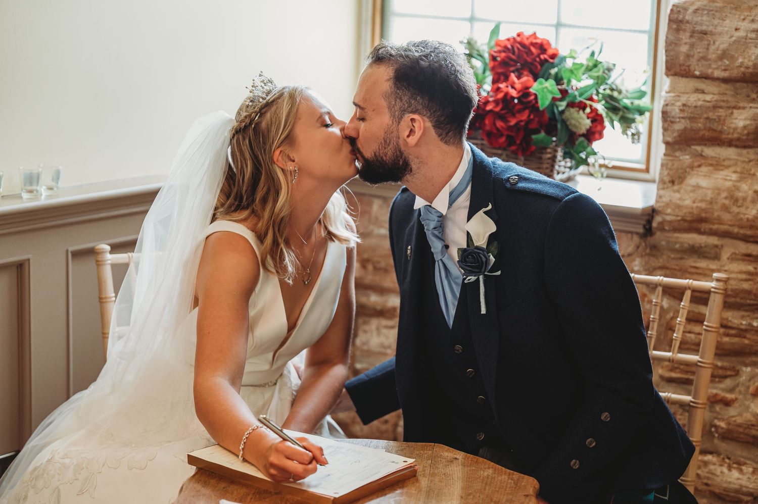 Newlyweds share a tender kiss at a rustic wooden table during their wedding ceremony.