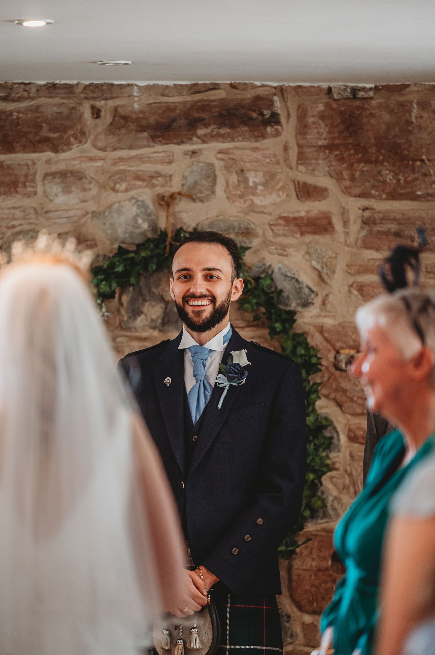 A Scottish wedding ceremony takes place in front of a rustic stone wall decorated with green foliage.