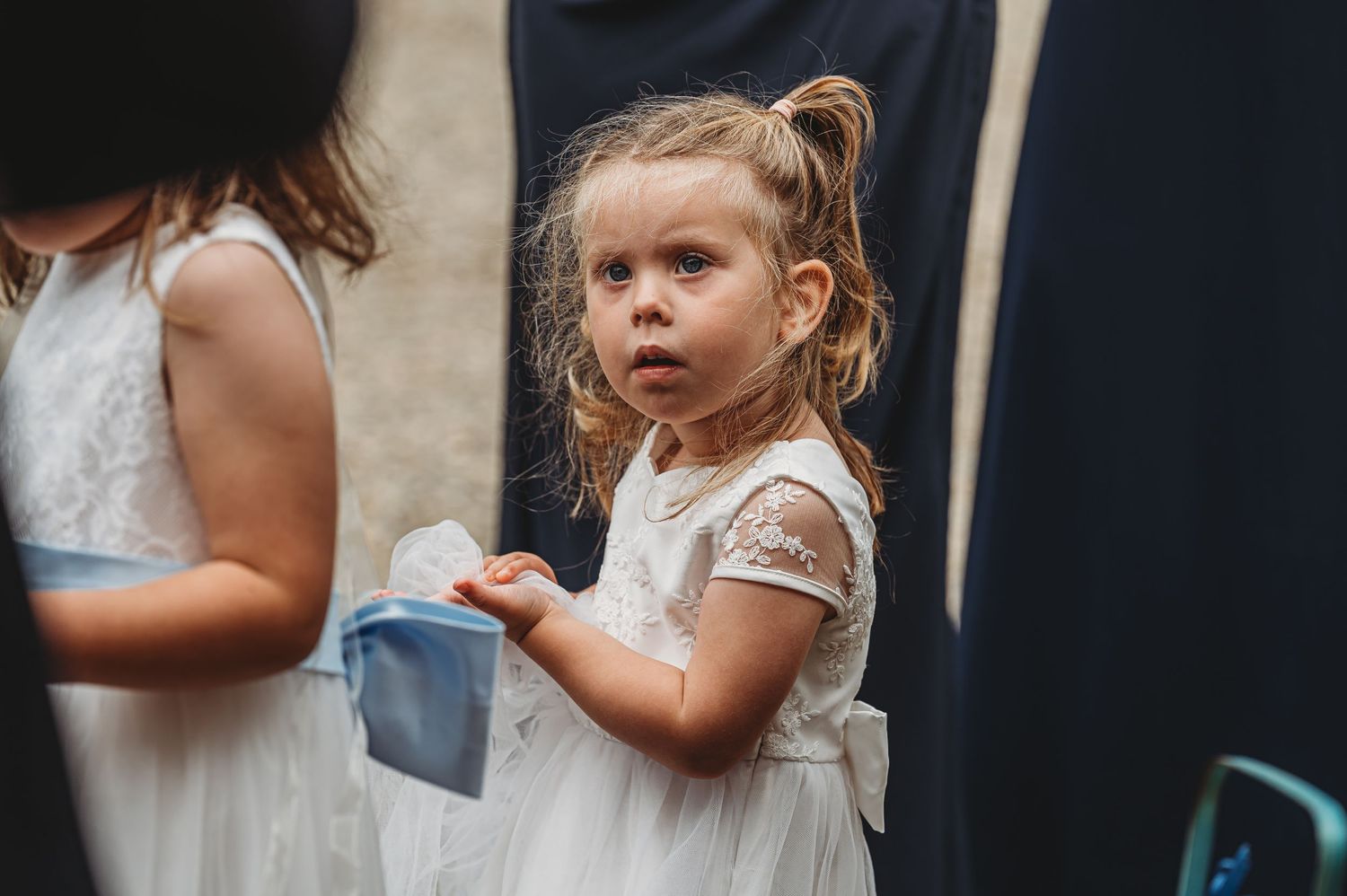 Small child in a white lace dress holds a blue cup at a formal event.