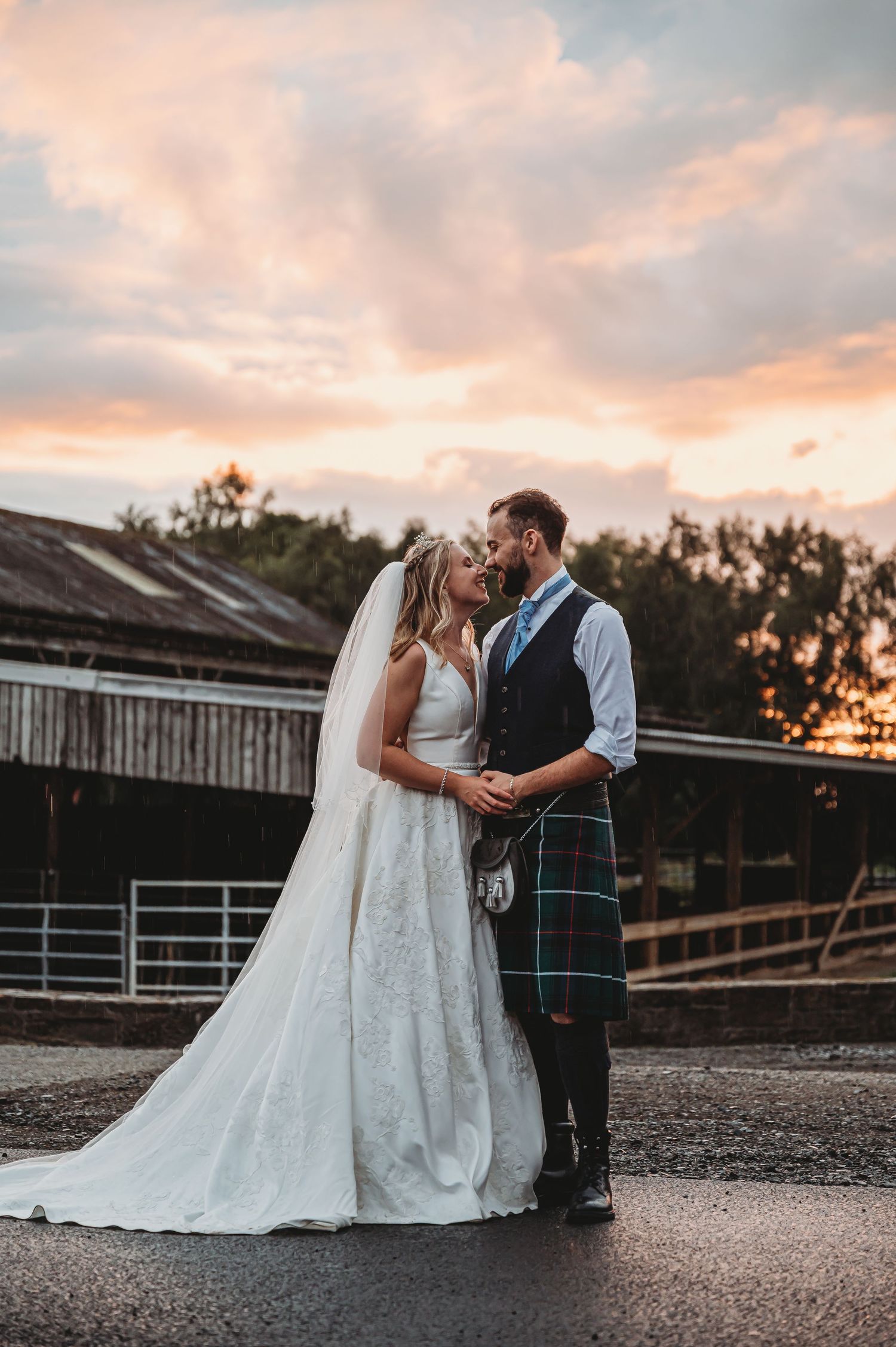 A romantic wedding couple in Scottish attire share an intimate moment at sunset outside a rustic barn building.