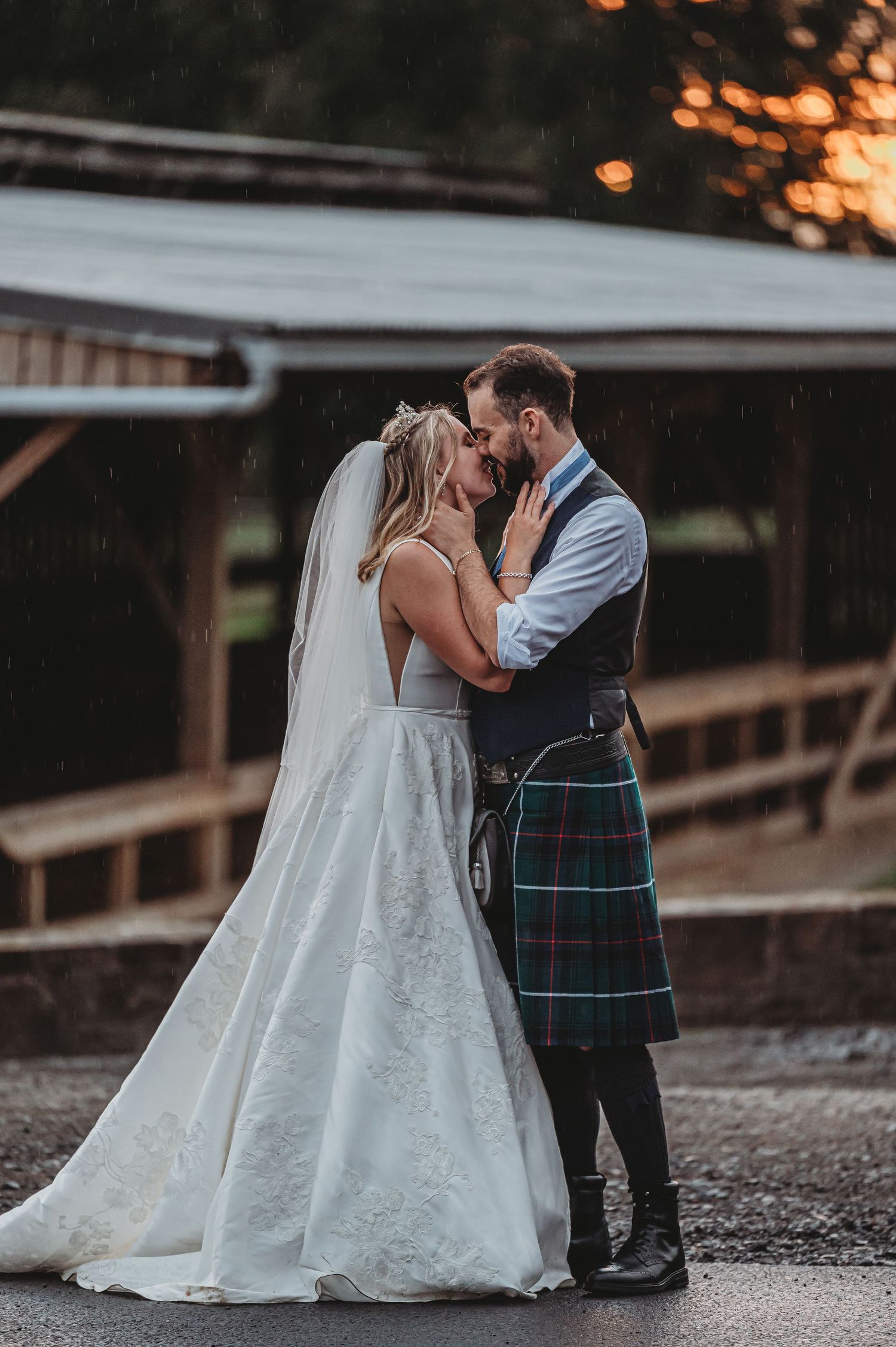 A romantic wedding couple embraces at sunset, with the groom wearing a traditional Scottish kilt and the bride in a flowing white gown.
