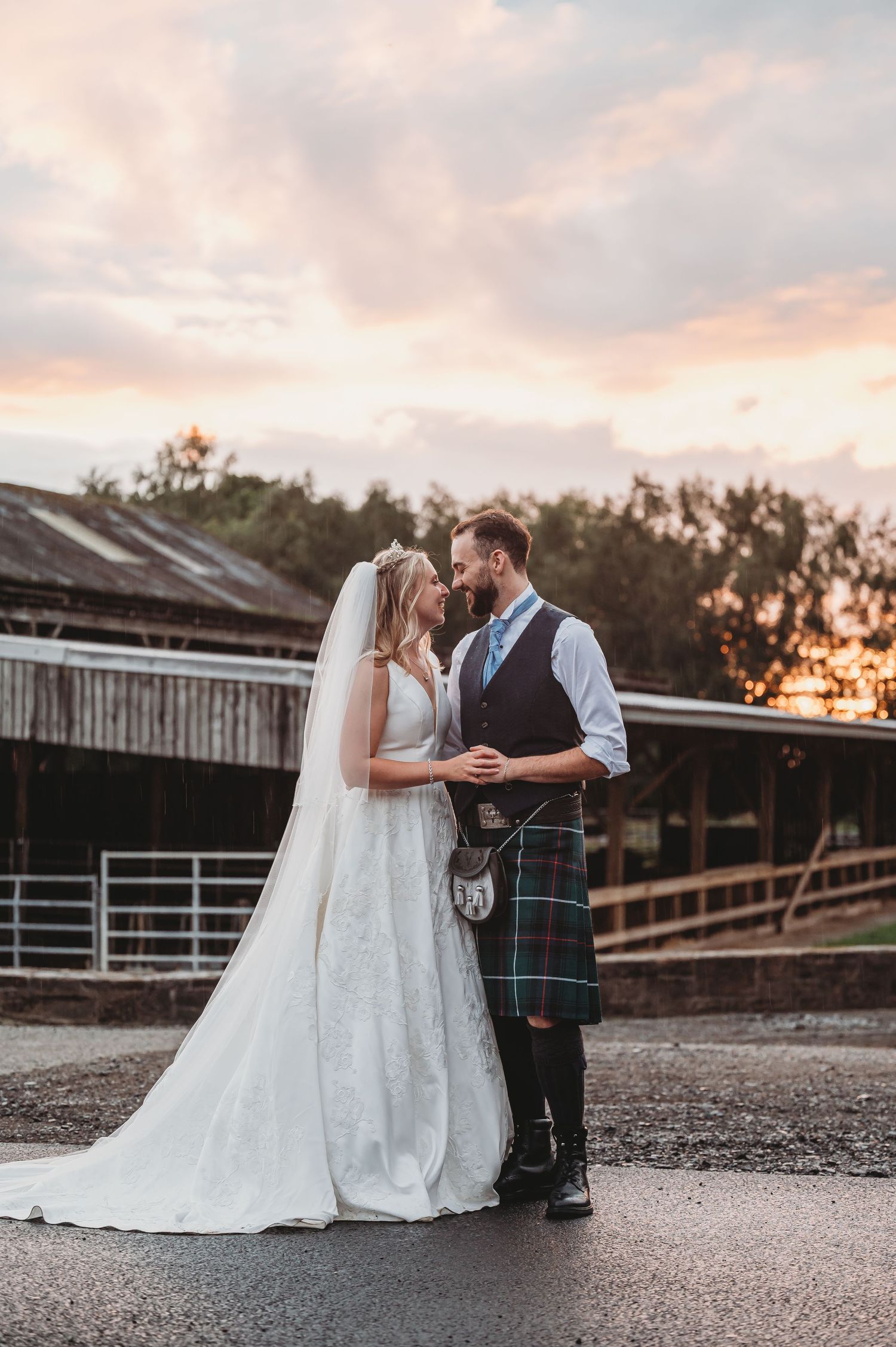 Couple shares romantic moment at sunset by rustic barn, with groom wearing traditional Scottish kilt and bride in white gown.