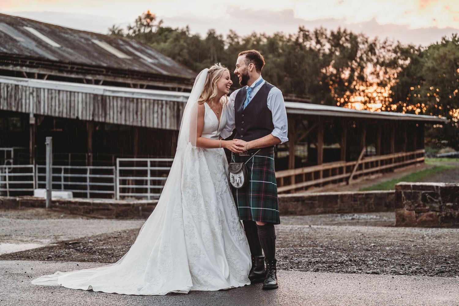 A couple in wedding attire embraces outside a rustic barn at sunset with a beautiful countryside backdrop.