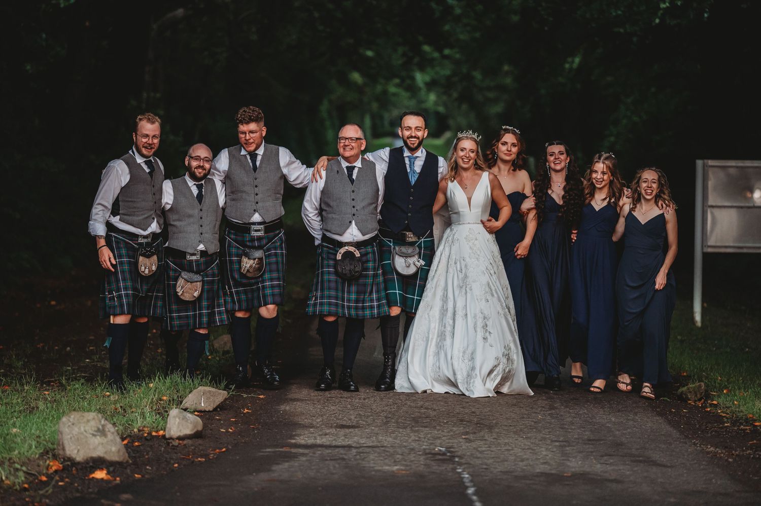 Wedding party poses on path with groomsmen in traditional kilts and bridesmaids in navy blue dresses.