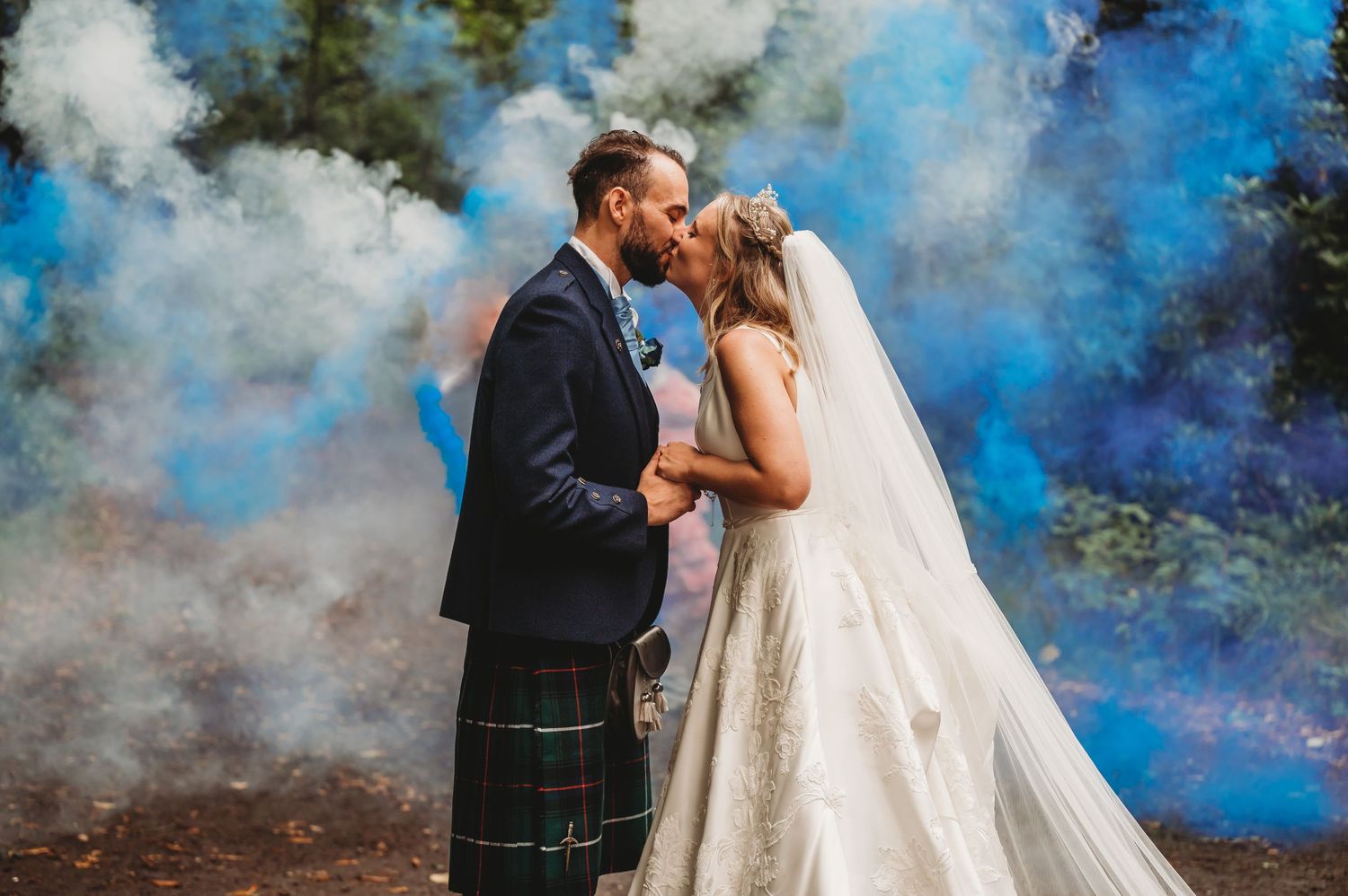 A bride and groom share a romantic moment surrounded by dramatic blue and white smoke in a forest setting.