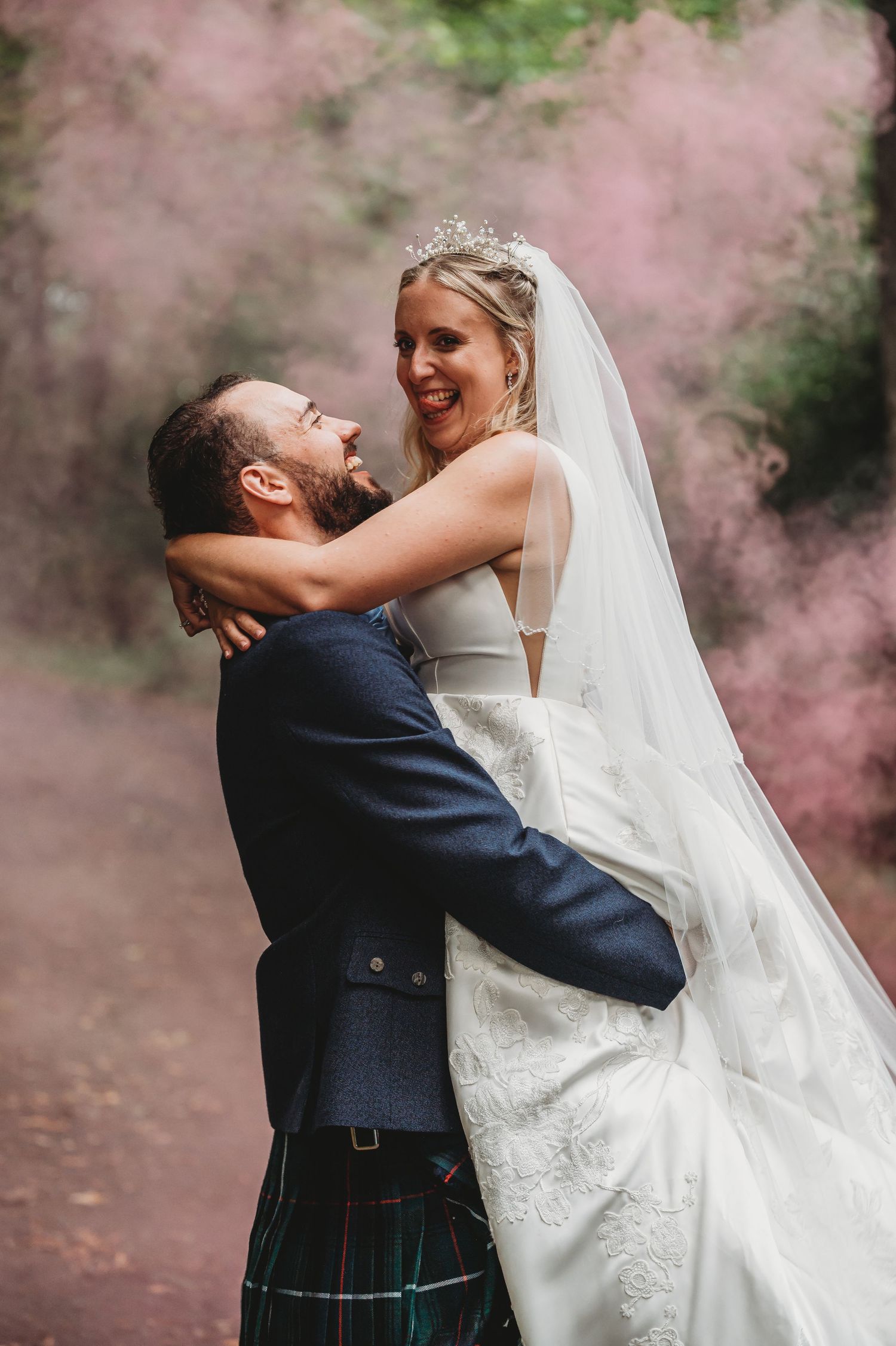 A joyful wedding couple embraces while surrounded by pink smoke in a romantic outdoor photo shoot.