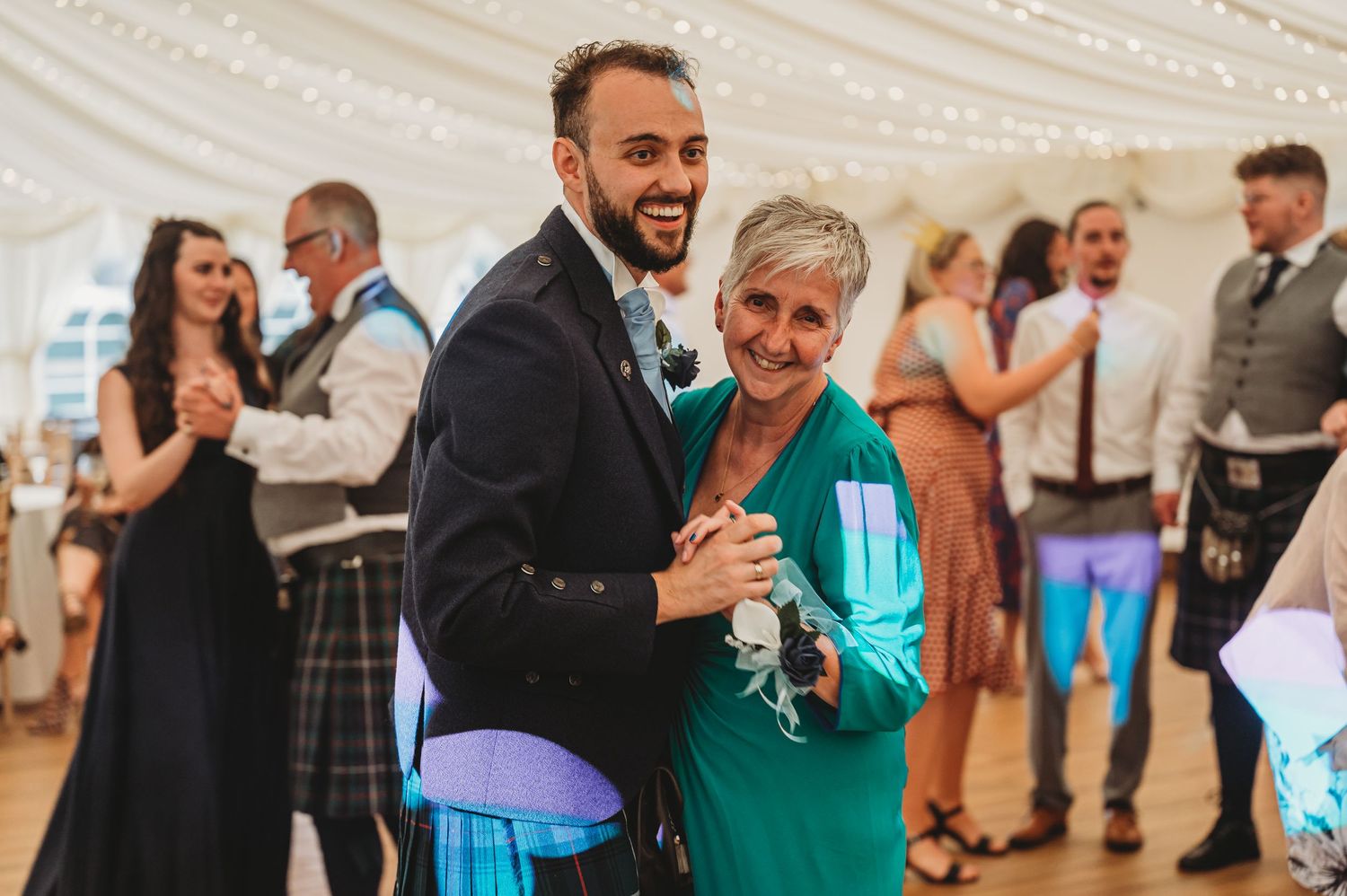 Wedding guests share laughs while dancing together at an elegant reception with blue ambient lighting.