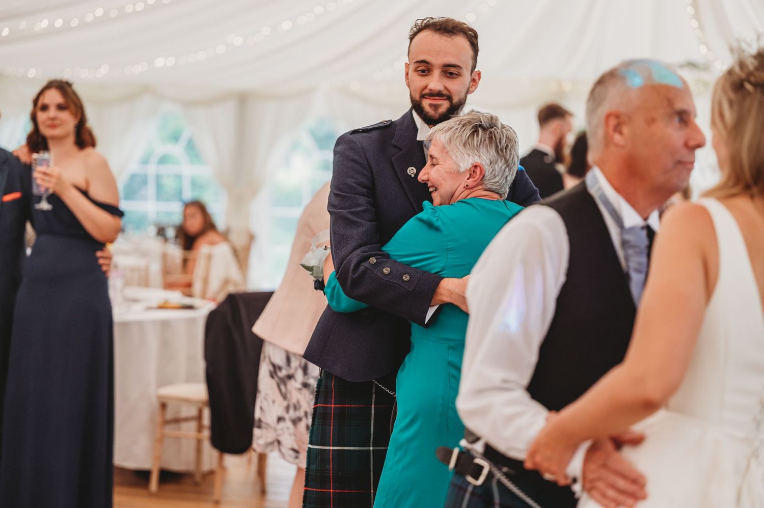 Guests share an emotional embrace during a wedding reception in a white tent venue.