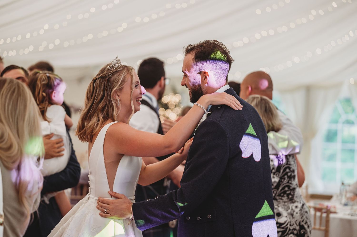 A couple dances close together under a wedding tent decorated with string lights while other guests dance around them.