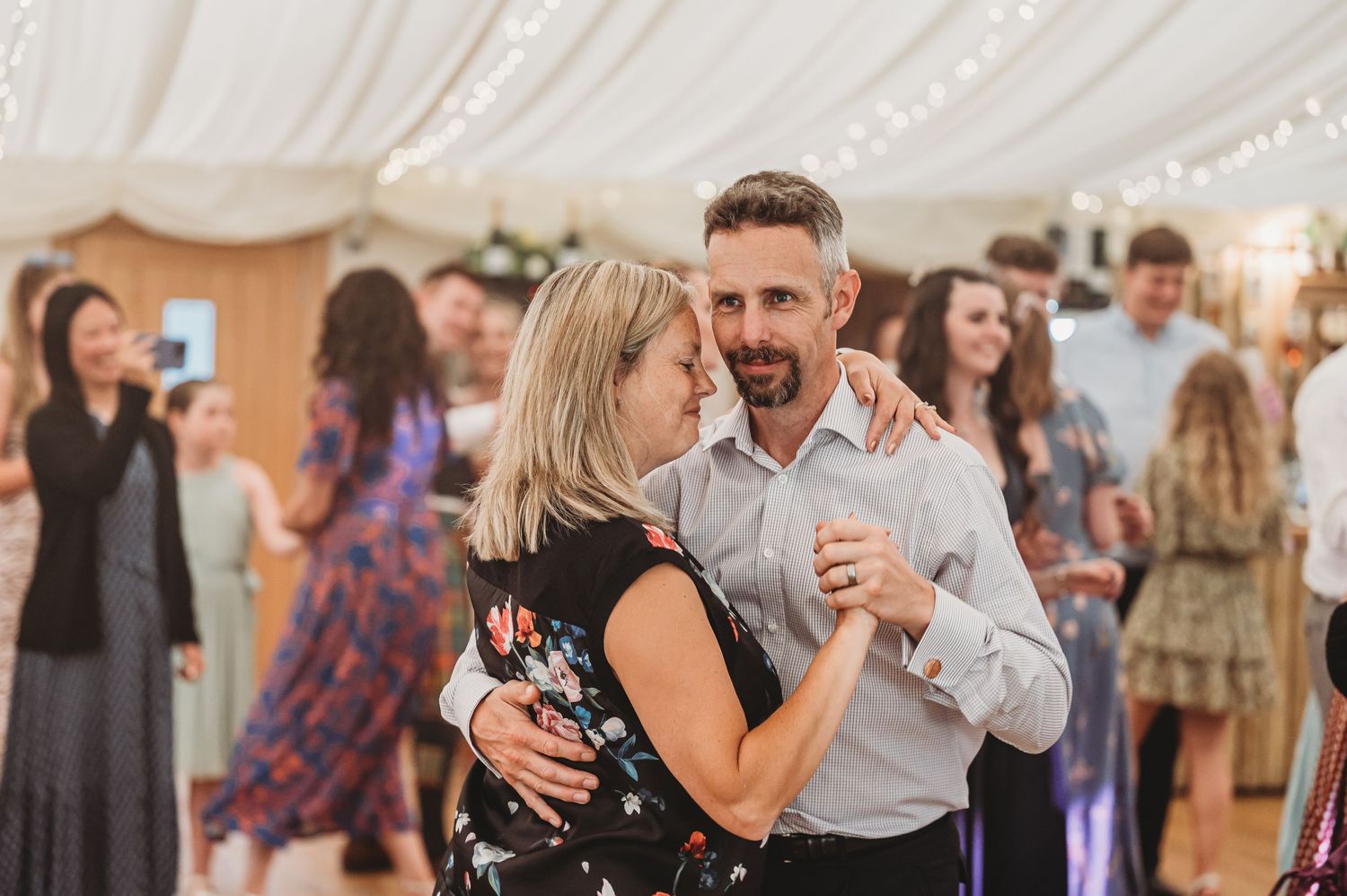 A couple slow dances together at a wedding reception under decorative string lights in an elegant venue.
