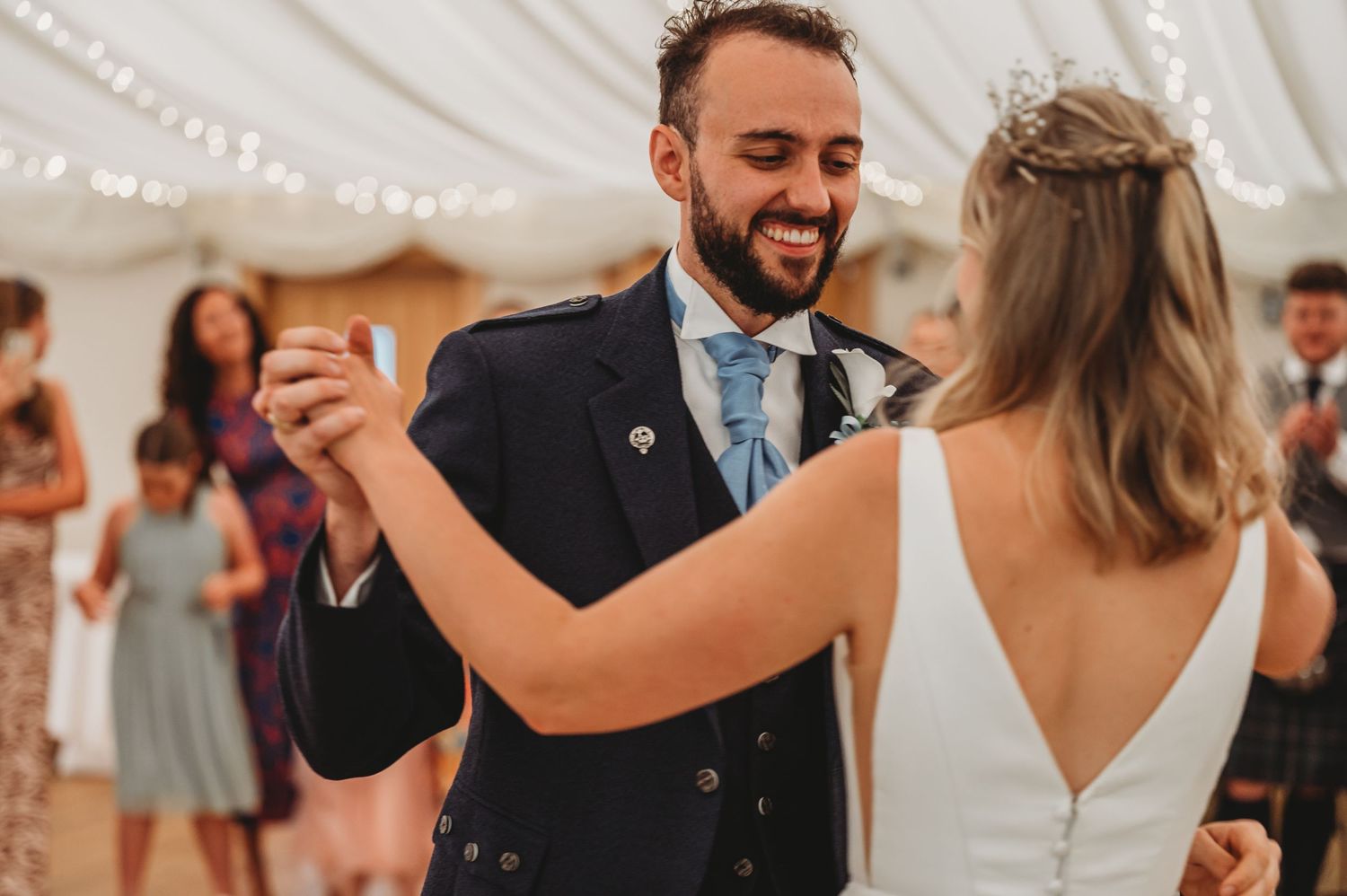 A bride and groom share their first dance at a wedding reception under white tent with smiling guests in the background.