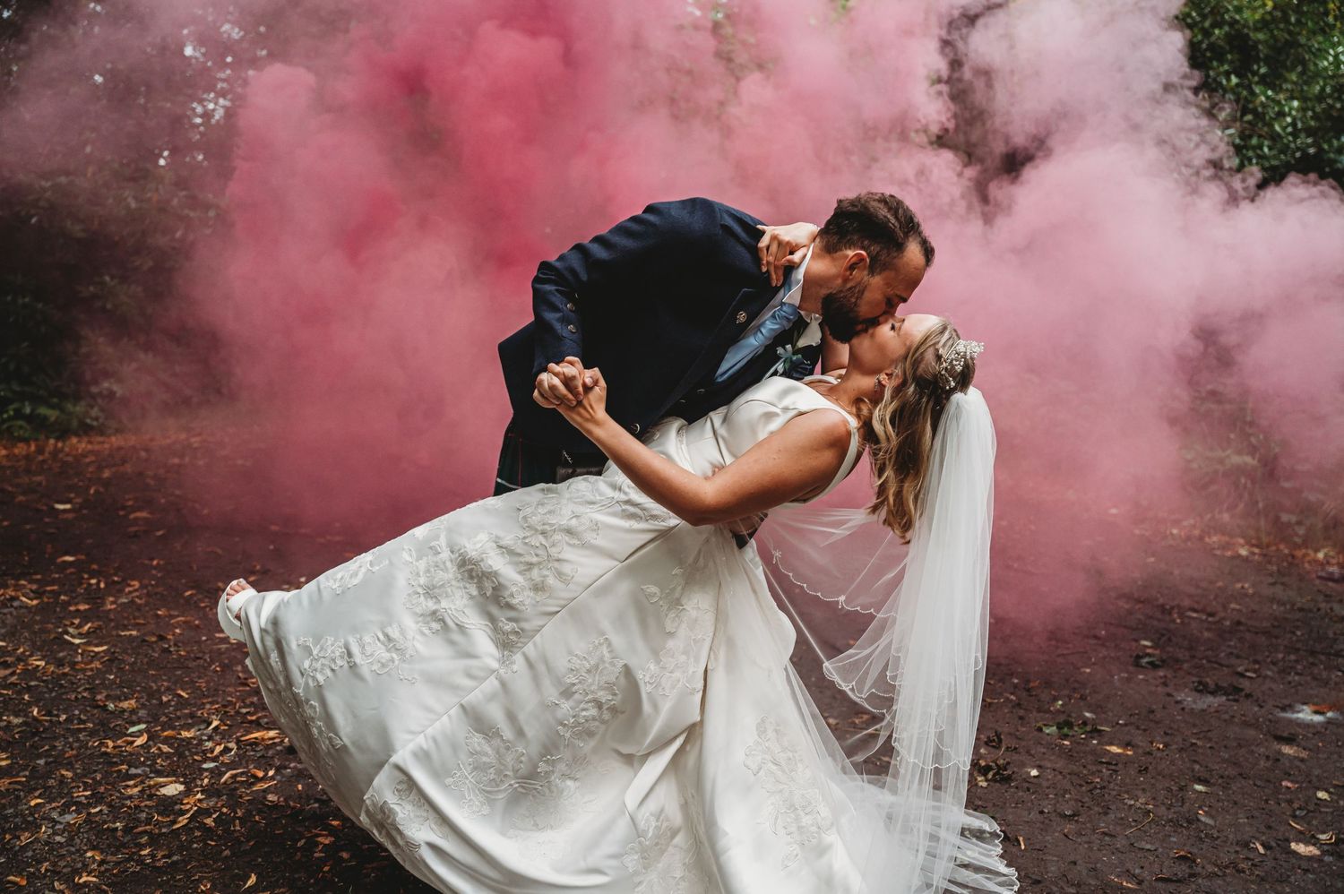 A romantic wedding couple shares a passionate dip kiss surrounded by dramatic pink smoke in a forest setting.