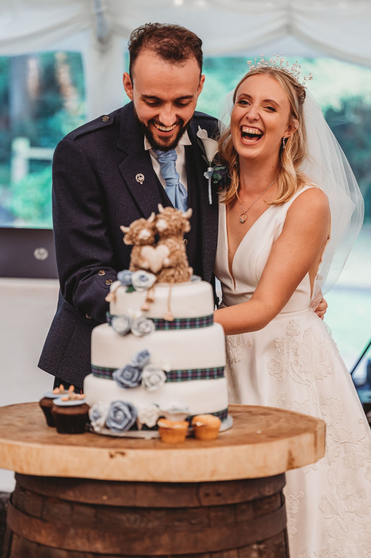 Happy couple laughing together at their wedding while cutting a beautiful three-tiered cake decorated with blue roses.