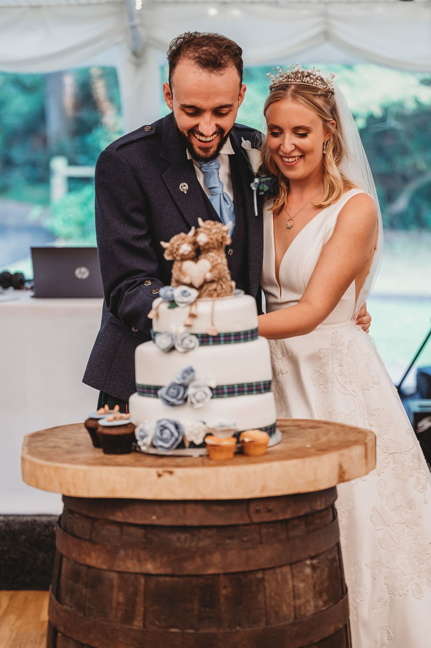 A happy couple cutting their elegant three-tiered wedding cake decorated with silver accents on a rustic wooden barrel.