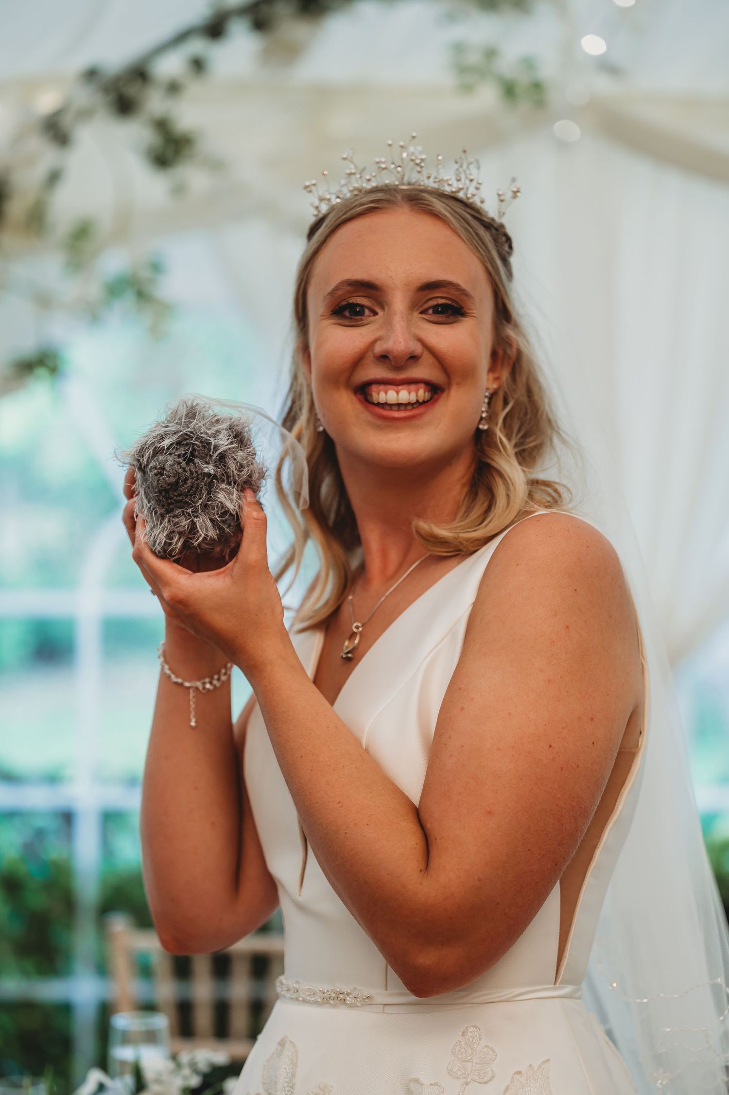 Bride in white wedding dress and crystal tiara smiles while holding a sparkling orb in festive indoor setting.