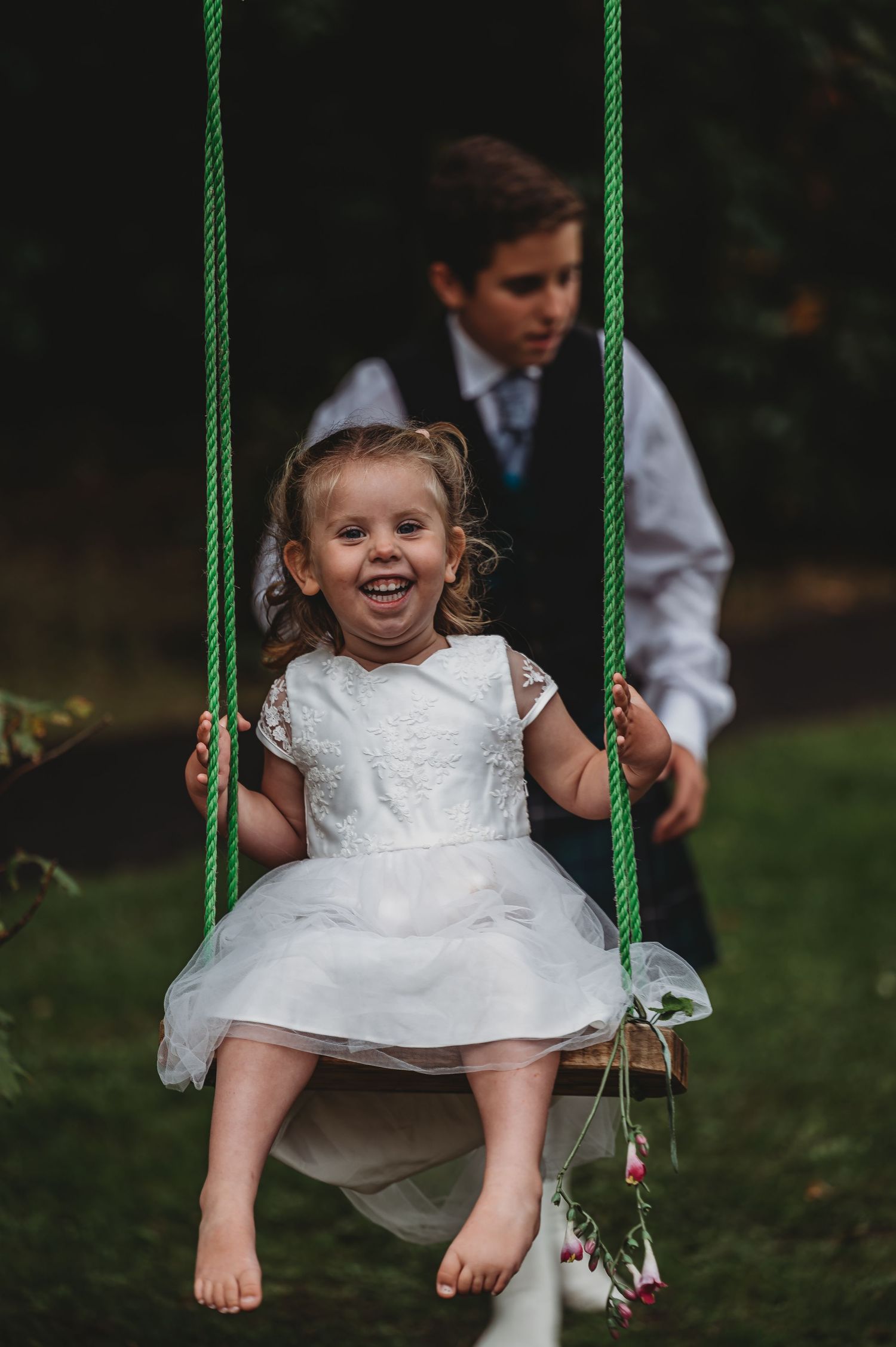 Joyful child in white dress swings on green rope swing in garden while being pushed from behind.
