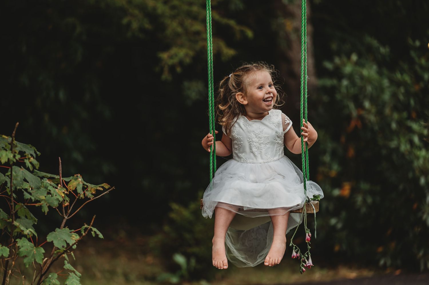 A child in a white dress sits on a green swing in a lush backyard garden setting.