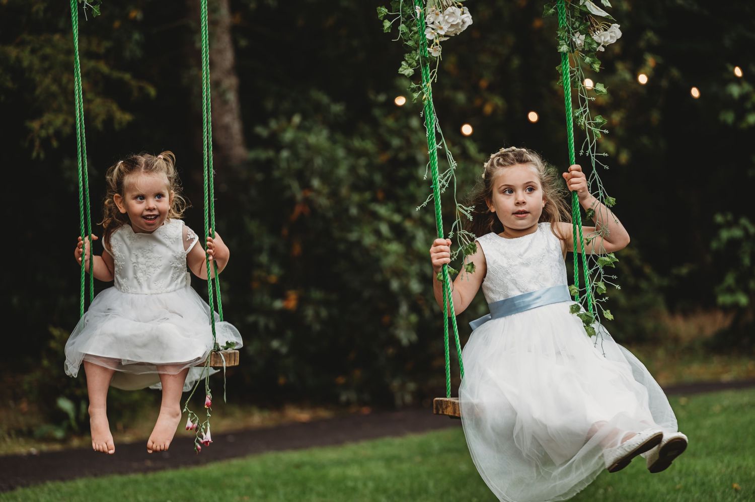 Two young children in white dresses swing together on green swings in a garden setting.