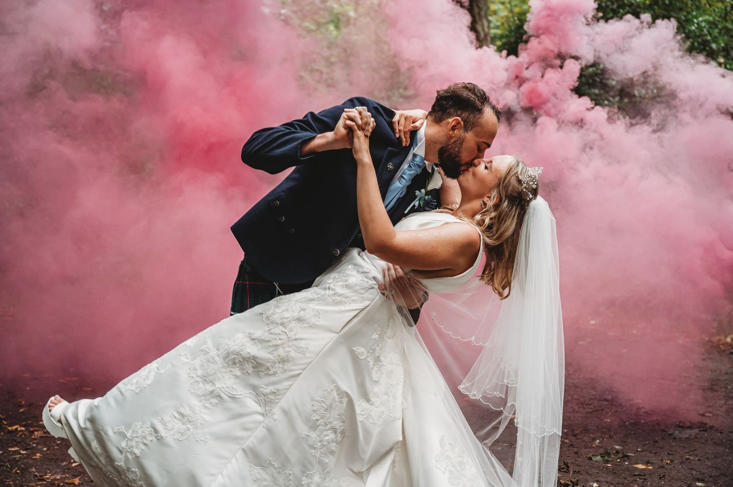 A romantic wedding couple embraces in a dramatic dip kiss surrounded by vibrant pink smoke bombs in an outdoor setting.