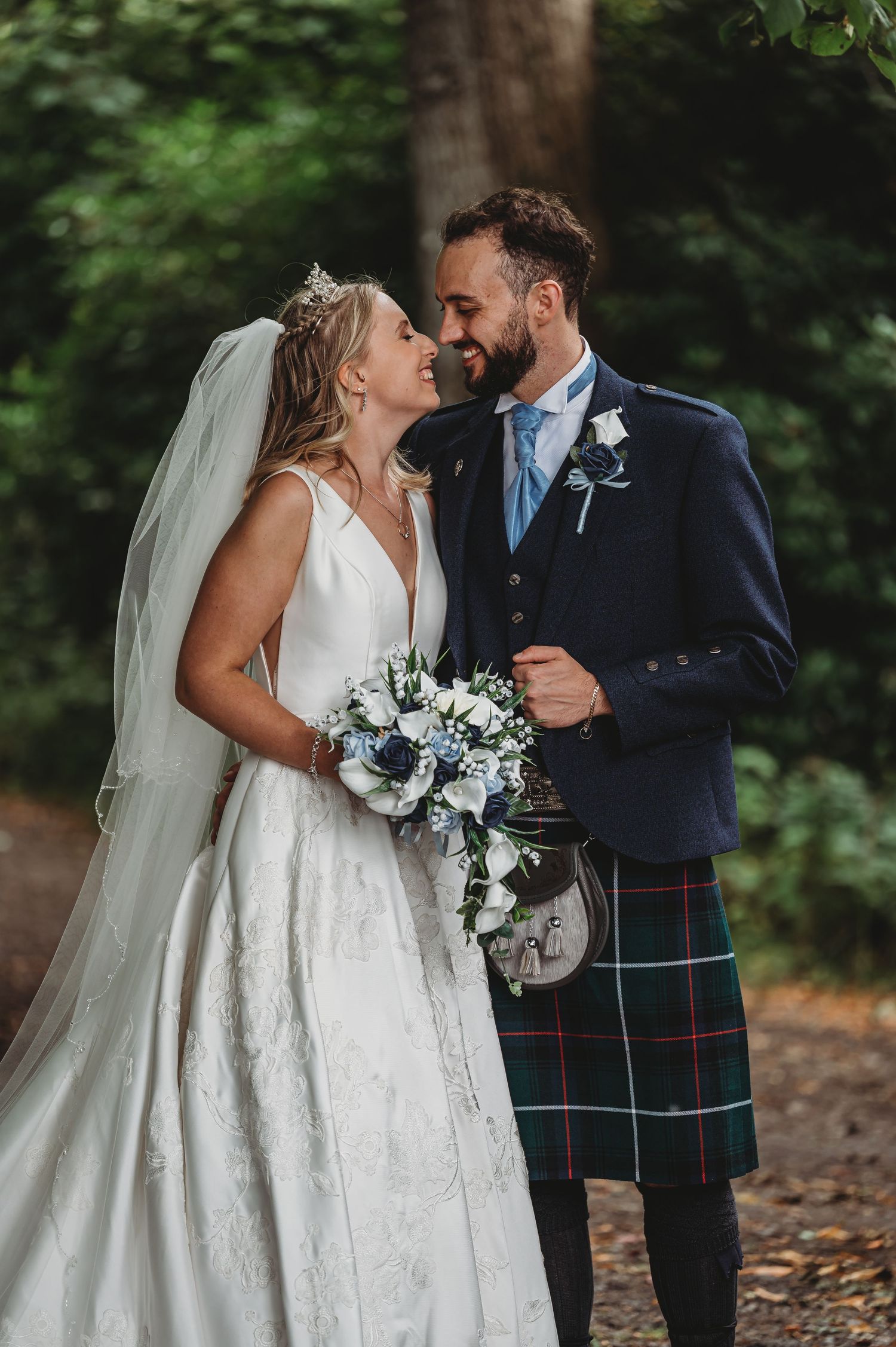 A couple shares an intimate moment in a forest setting, with the groom in traditional Scottish kilt and bride in white gown.