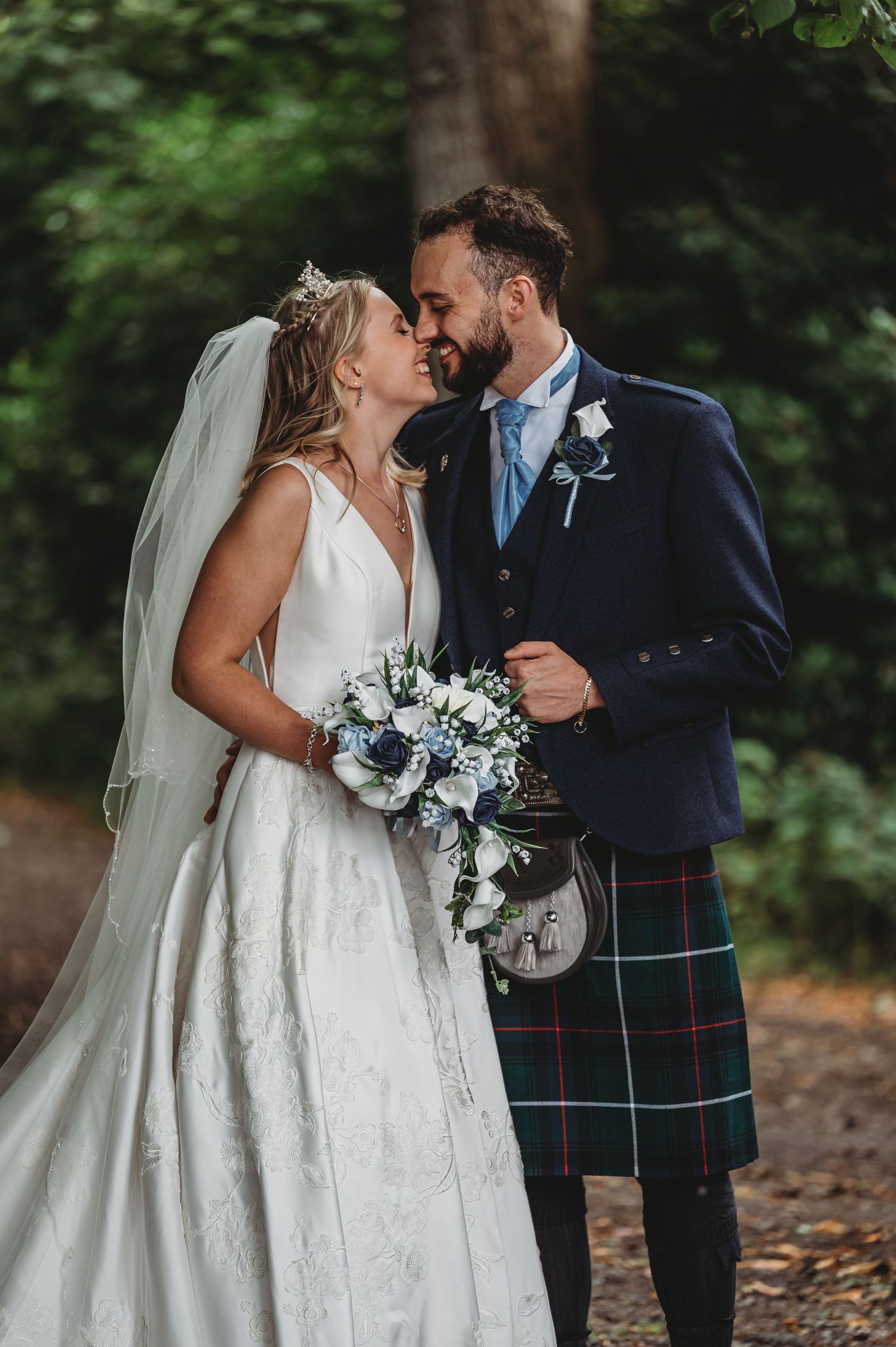 A bride in a white dress and groom in a traditional kilt share a romantic moment in a wooded setting.